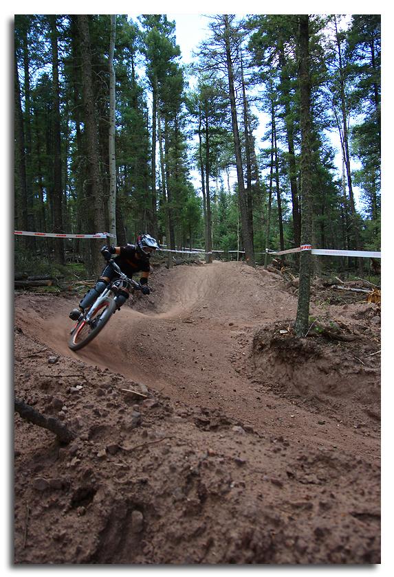 Mountain biker navigating a dirt trail in a forest, leaning into a turn with trees in the background. Angel Fire Bike Park mountain bike trail.