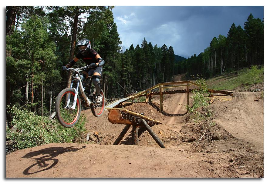 A mountain biker in a helmet and protective gear jumps off a dirt ramp, with a wooden structure and a forest of trees in the background. The scene captures a moment of action and adventure in a natural outdoor setting. Angel Fire Bike Park mountain bike trail.