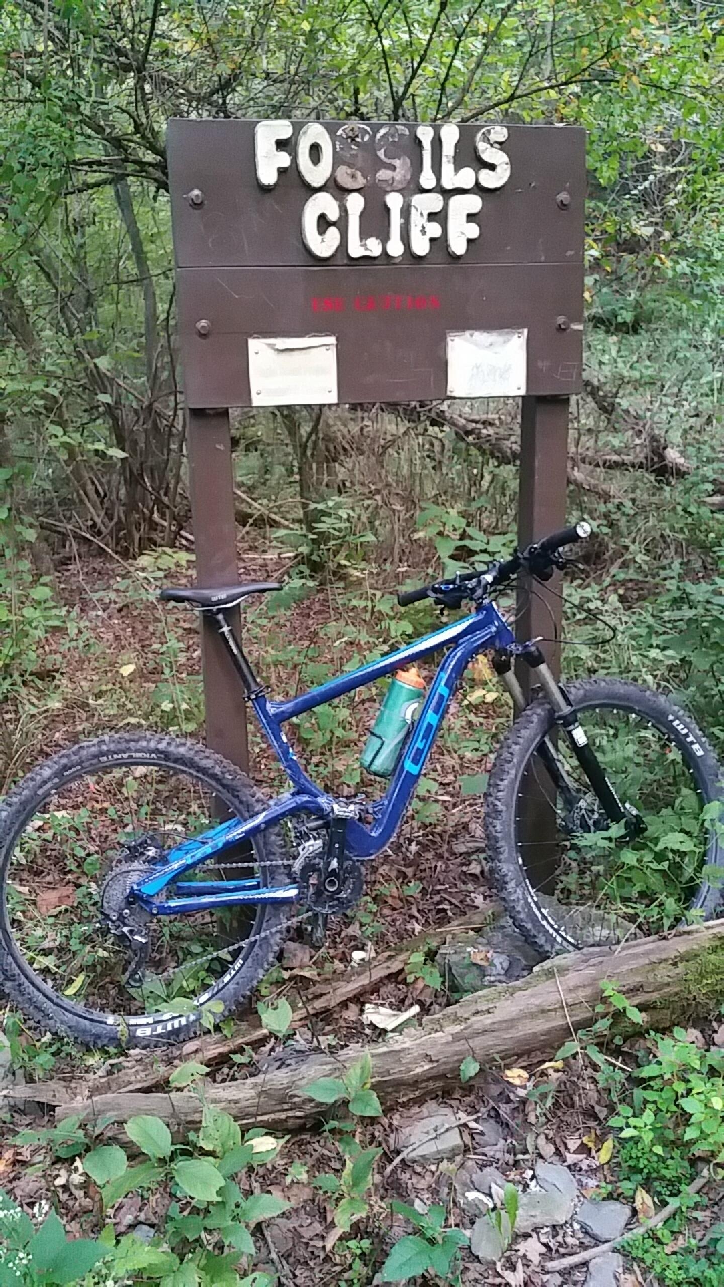 GT Helion: A blue mountain bike leans against a sign that reads "Fossils Cliff" in a forested area. The ground is covered with fallen leaves and small plants, and the background features dense greenery and trees.