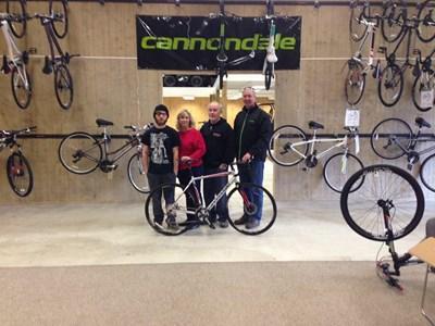 A group of three people poses with a bicycle in a bike shop, which features several hanging bicycles on the walls. The background includes a prominent Cannondale sign.