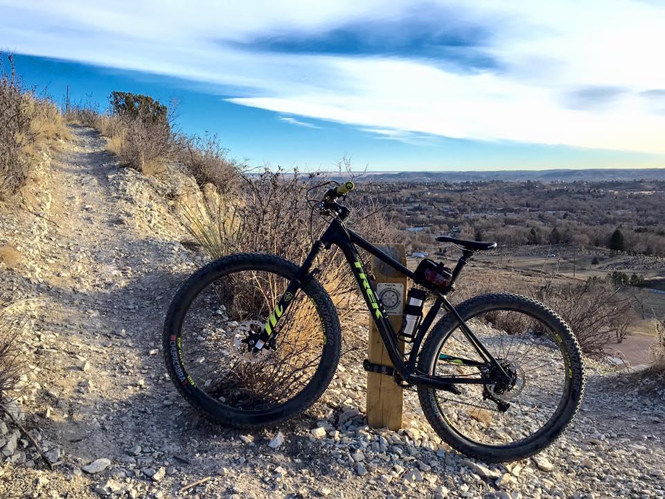 Trek Stache 9: A mountain bike resting on a scenic gravel trail surrounded by sparse vegetation. In the background, a panoramic view stretches over a valley with a clear sky, showcasing the landscape and distant hills.