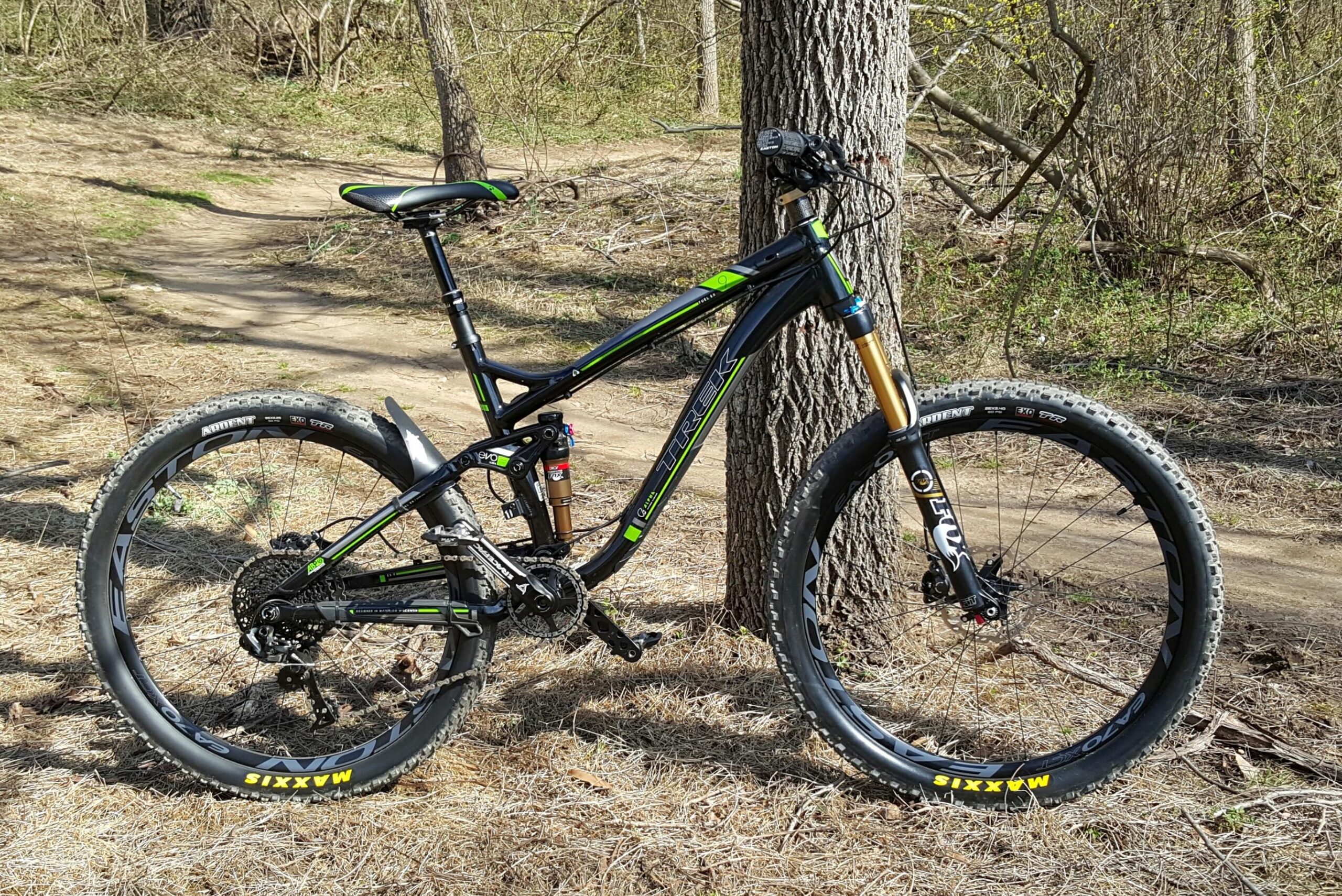 Trek Fuel EX 9: A black and green mountain bike stands against a tree in a grassy area with a dirt path visible in the background. The bike features thick tires designed for off-road terrain, a suspension system, and a sleek frame, indicating it's built for rugged trails and outdoor adventure.