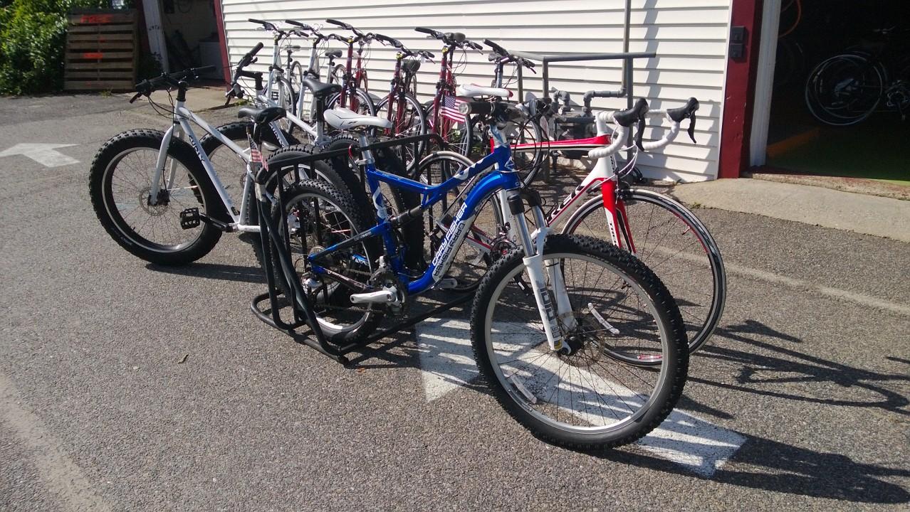 A row of bicycles parked in a bike rack outside a shop, featuring various types including road bikes and mountain bikes, with a few having bright colors like blue and white. The background shows a part of the shop with a red door and greenery.