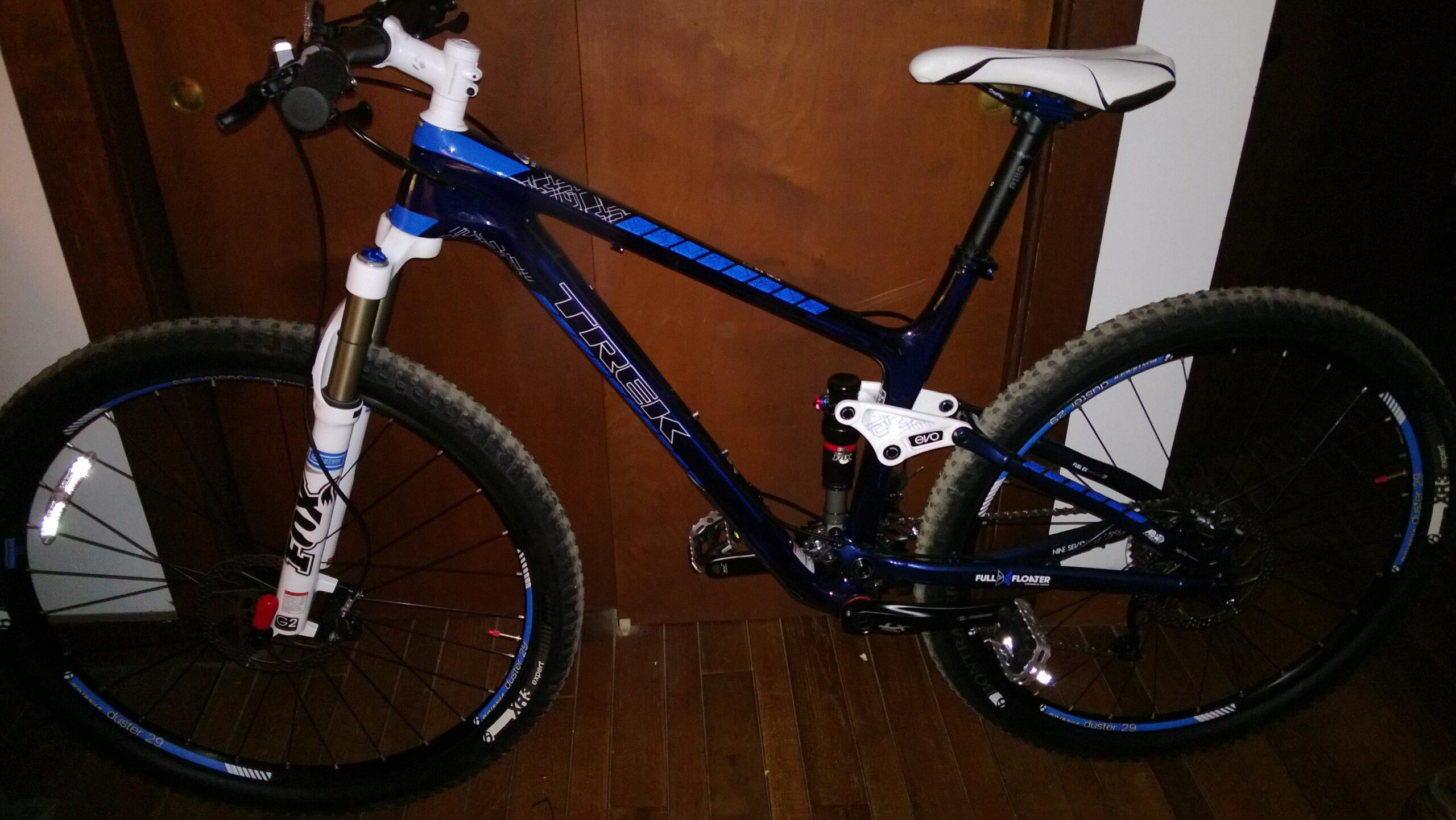 Trek Fuel EX 9.7 29: A blue Trek mountain bike leaning against a wooden wall. The bike features a white and blue saddle, thick tires, and a suspension system. The background shows a closed door with wooden panels.