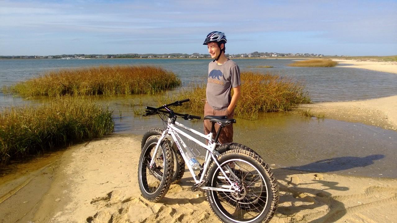 Surly Pugsley: A young man wearing a helmet stands next to a mountain bike on a sandy beach near a body of water. In the background, there are patches of tall grass and a clear blue sky. The scene conveys a relaxed outdoor atmosphere, perfect for biking and enjoying nature.