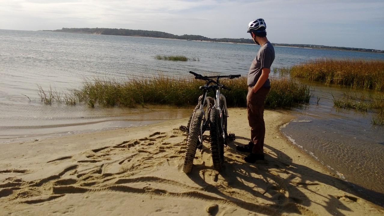 Surly Pugsley: A person standing on a sandy beach beside two mountain bikes, looking out over a calm body of water with grassy vegetation along the shore and a distant landmass visible in the background under a partly cloudy sky.