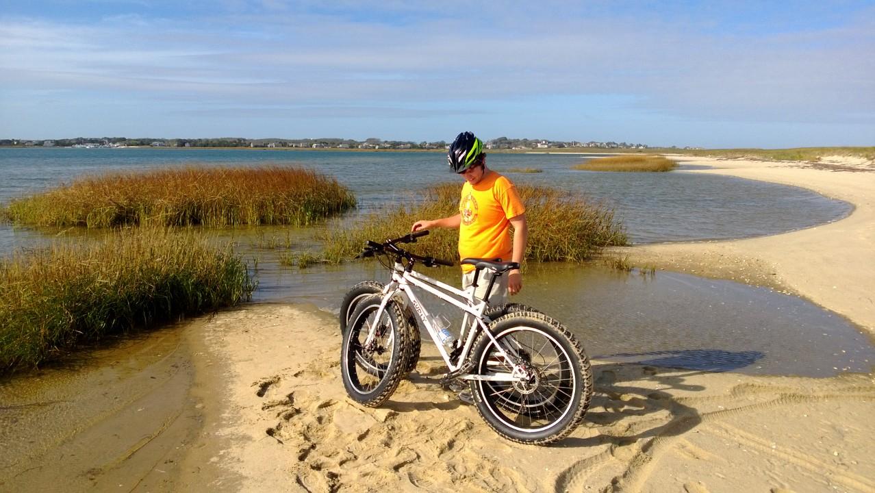 Surly Pugsley: A person in an orange shirt and a helmet standing next to a fat-tire bike on a sandy beach near a body of water, with grass and distant homes visible in the background.