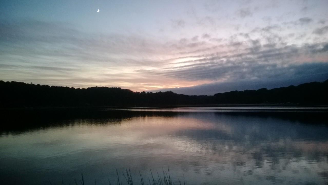 Serene lake at sunset with soft ripples reflecting the colorful sky, featuring shades of pink and purple, and a crescent moon in the clear sky above. The surrounding landscape includes silhouette outlines of trees on the horizon. Volunteer Park/Chatham Ponds mountain bike trail.