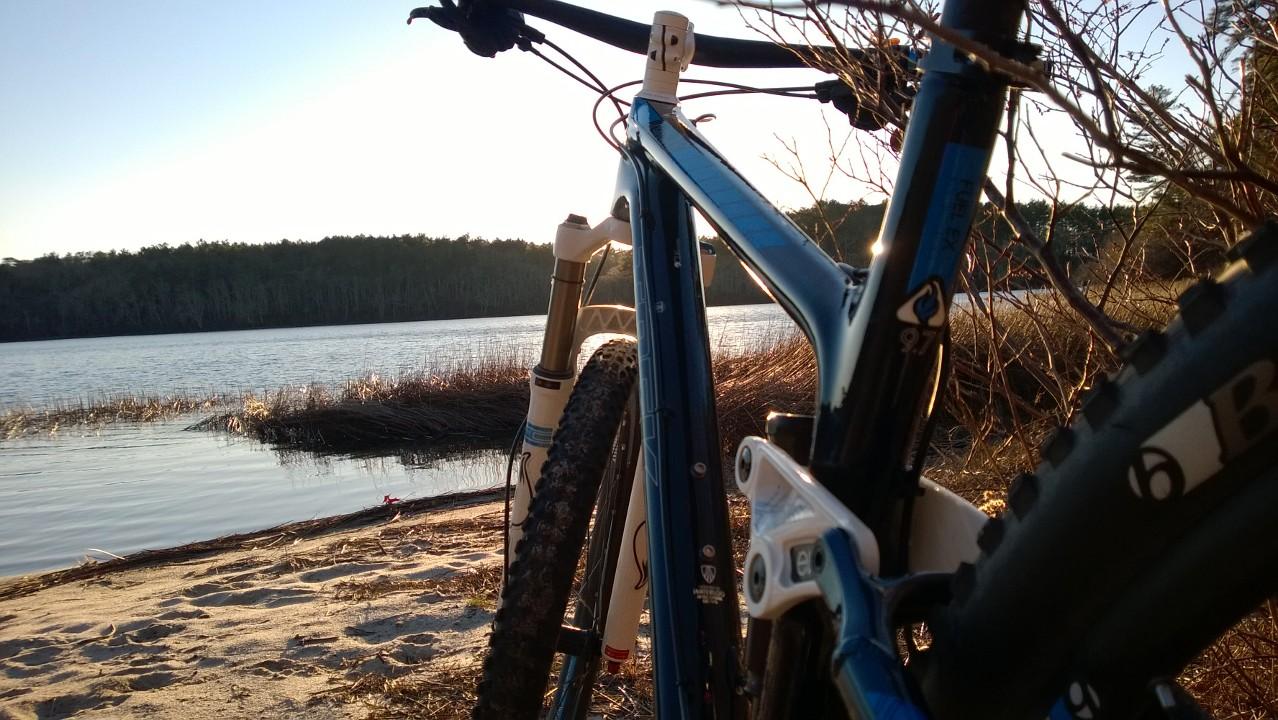Trek Fuel EX 9.7 29: Two mountain bikes are positioned on a sandy lakeshore, with a serene body of water reflecting the sunset in the background. Tall grasses and trees line the opposite shore, creating a tranquil outdoor scene.