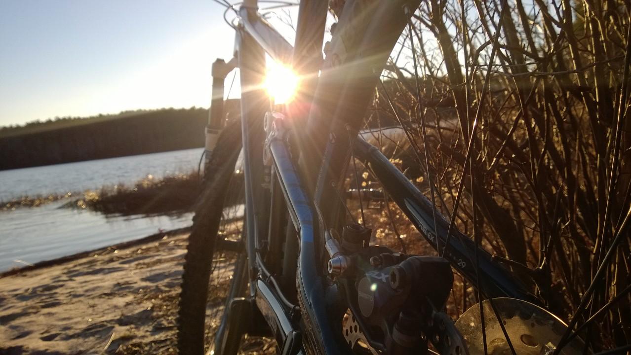 Trek Fuel EX 9.7 29: A close-up view of a bicycle resting near a body of water at sunset. The sun shines brightly through the bike's frame, creating a starburst effect, while shrubs and grasses surround the scene. The calm water reflects the warm colors of the sunset, enhancing the peaceful atmosphere.