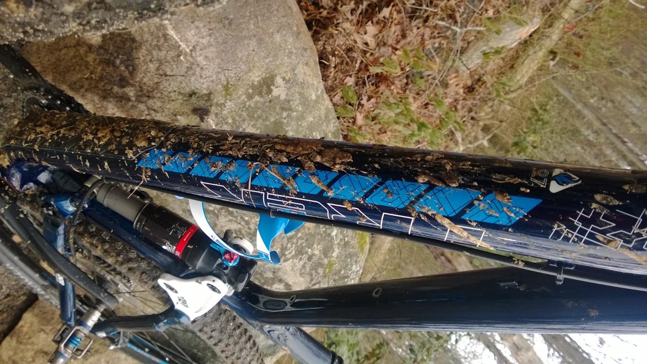 Trek Fuel EX 9.7 29: A close-up view of a mountain bike frame covered in mud, with a noticeable blue design on the top tube. The bike is positioned on rocky terrain, showcasing its rugged use. Leaf litter can be seen in the background, indicating a natural outdoor setting.