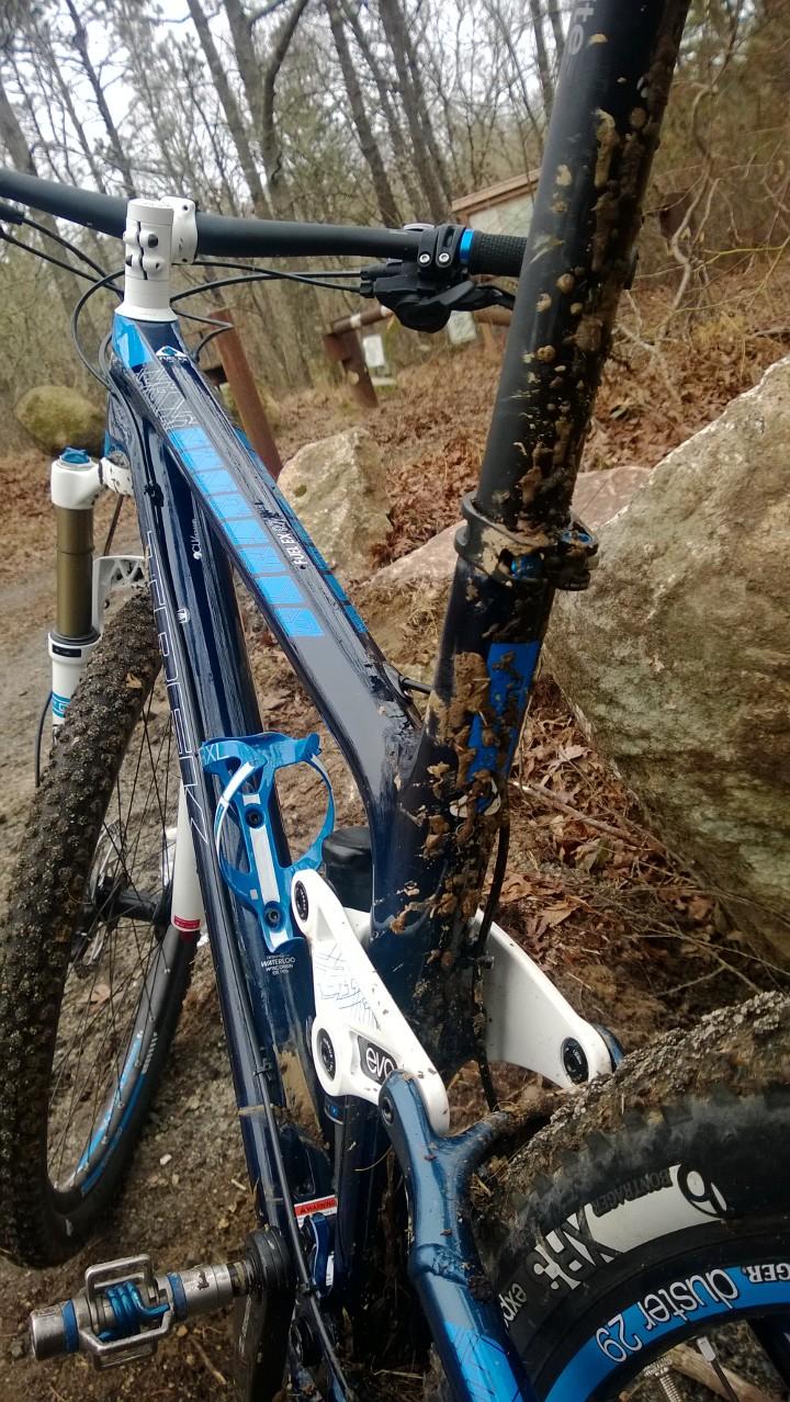 Trek Fuel EX 9.7 29: A close-up view of a mountain bike covered in mud, showcasing details such as the frame, handlebars, and drivetrain. The bike is parked on a dirt path surrounded by trees and rocks, suggesting a recent off-road ride. The muddy splashes indicate heavy use, highlighting the bike's adventurous terrain capability.
