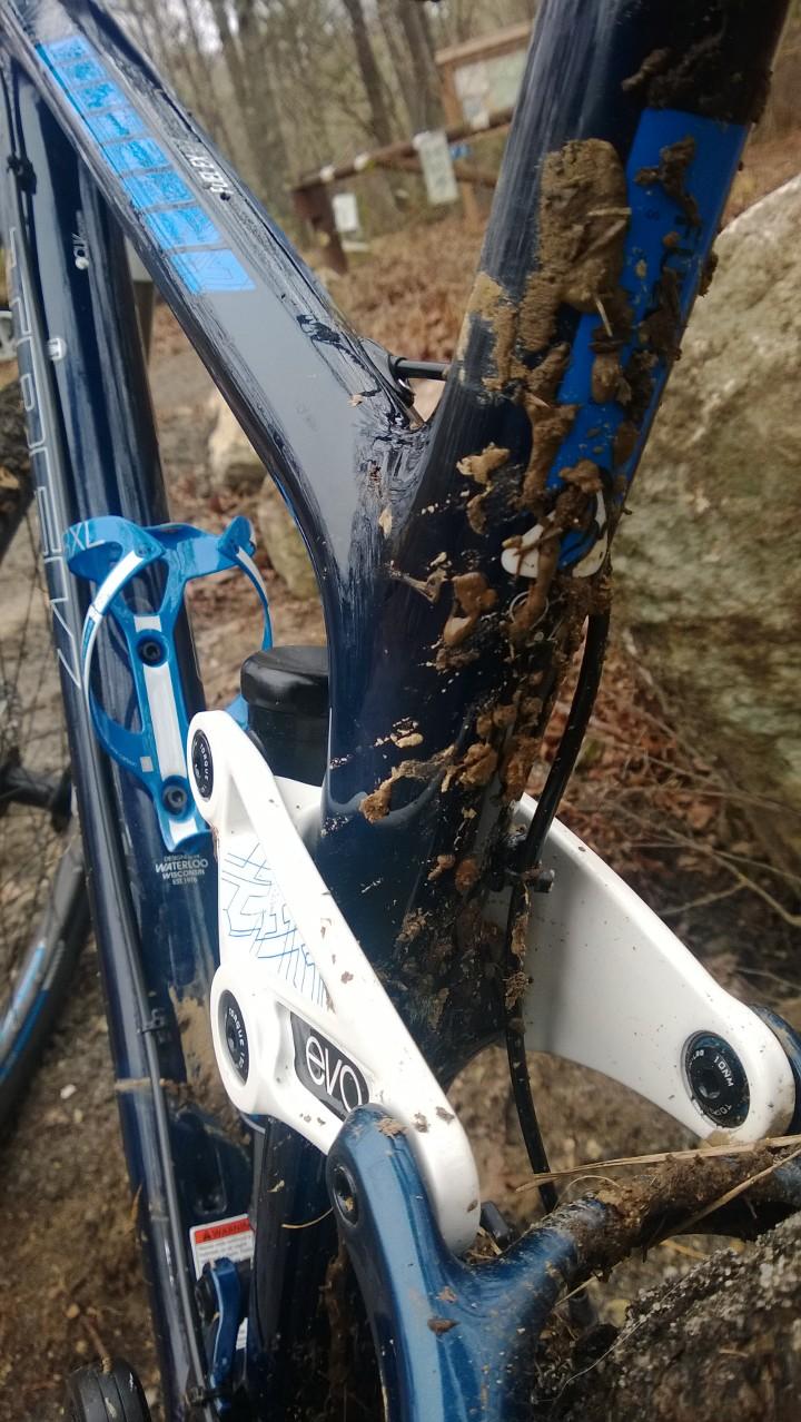 Trek Fuel EX 9.7 29: A close-up of a muddy bicycle frame, showcasing a blue and white mountain bike. The frame is splattered with dirt, particularly around the rear triangle and shock area. In the background, a blurred signpost and fallen leaves suggest a natural outdoor setting.