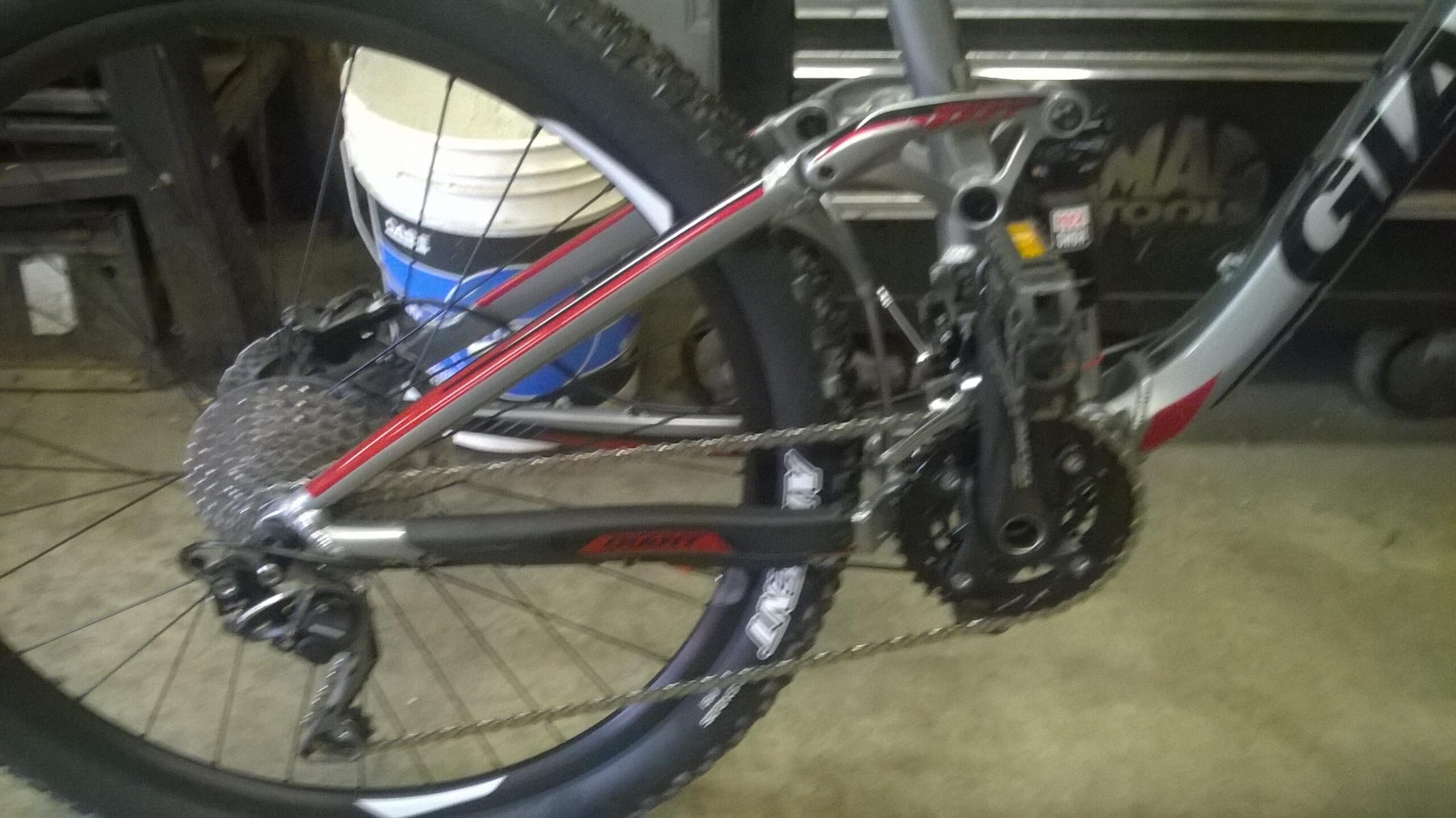 Giant Trance 27.5 3: Alt text: Close-up view of the rear derailleur and drivetrain of a mountain bike, featuring a silver and black frame with red accents. The bike is positioned in a workshop setting with a blurred background of tools and containers.