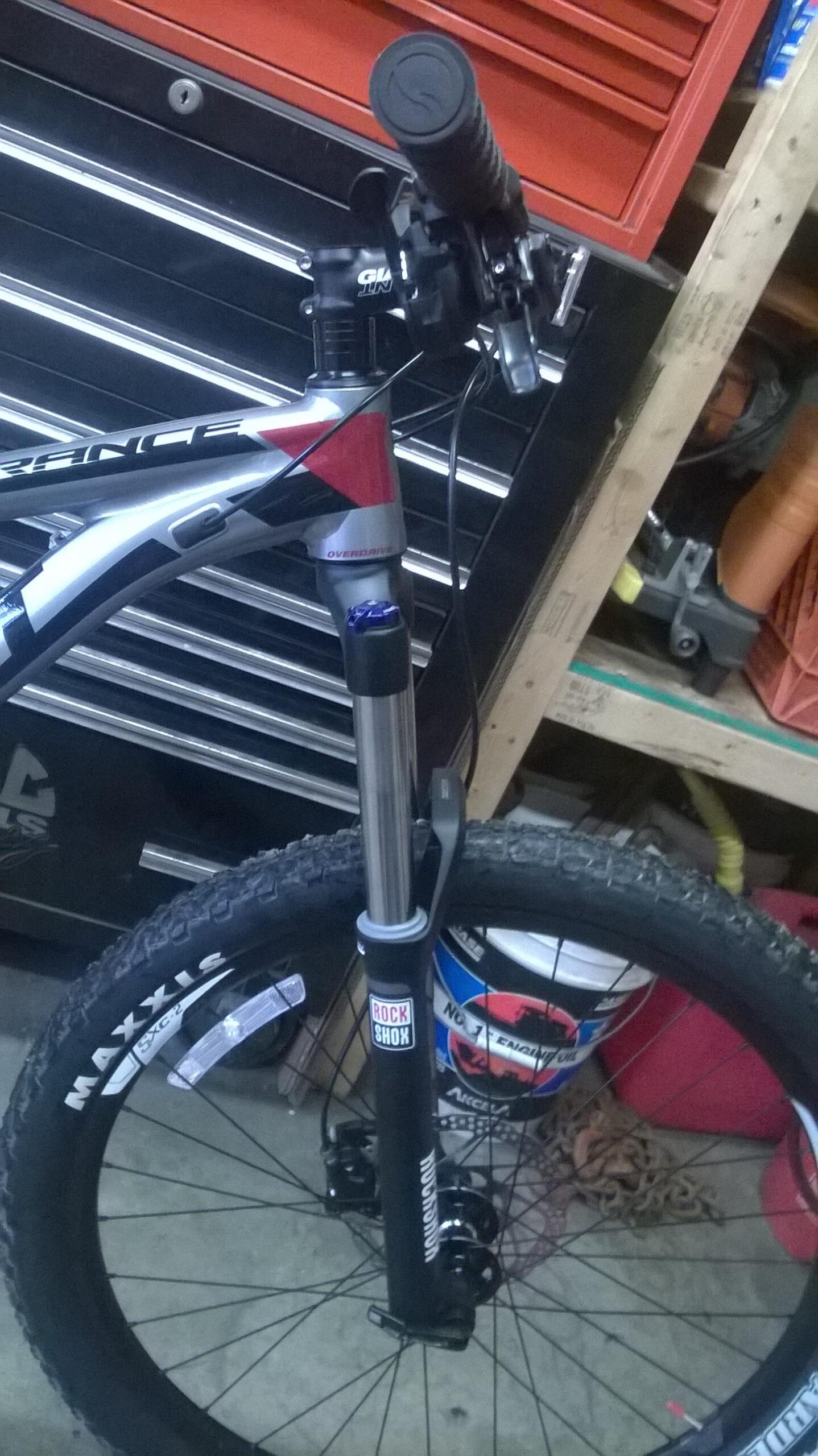Giant Trance 27.5 3: Image of a mountain bike with a close-up view of the front fork, handlebars, and tire. The bike features a silver and red frame, with a black suspension fork branded "Rock Shox" and a "Maxxis" tire. The background includes a toolbox and various items, indicating a workshop setting.