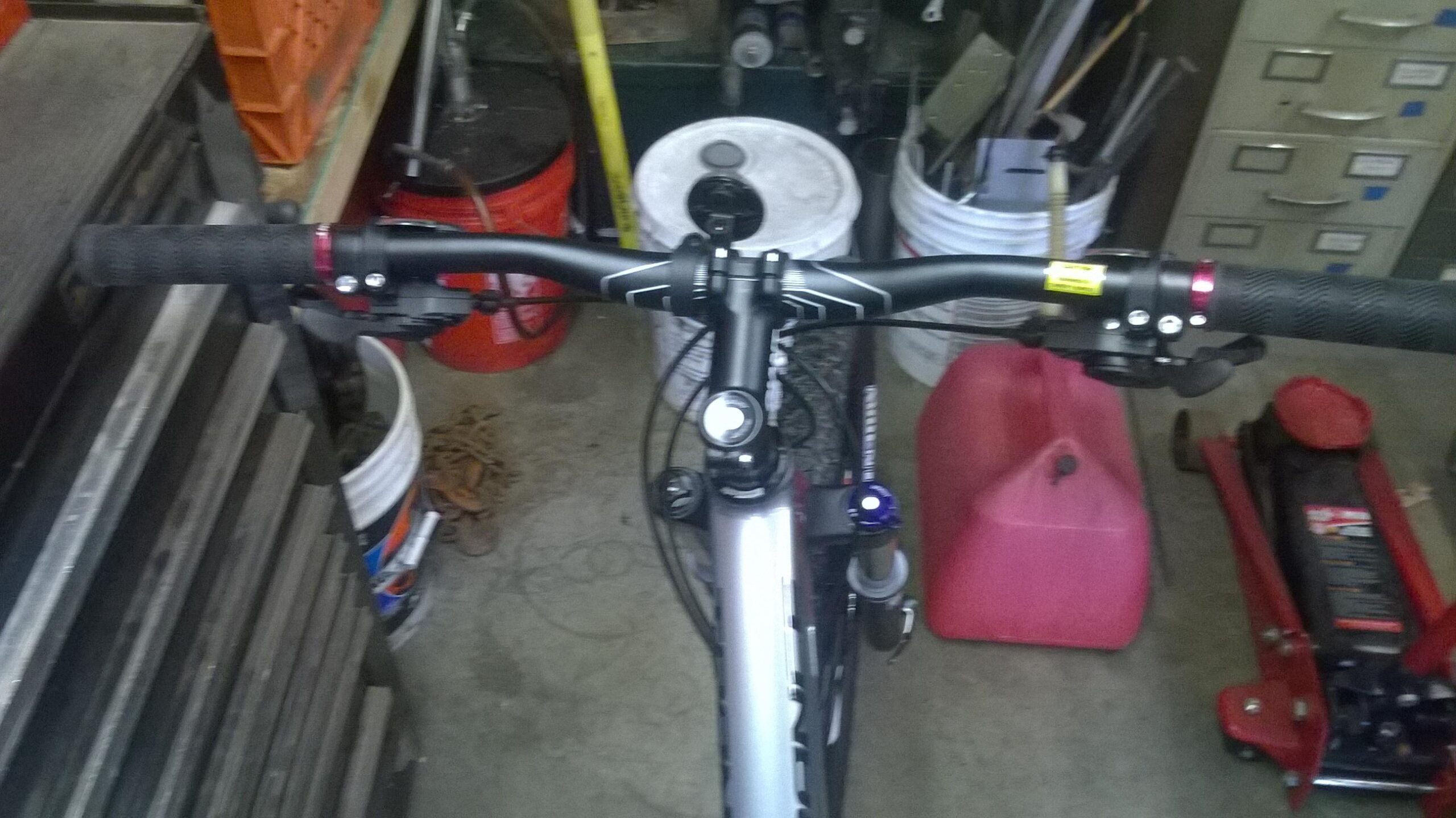 Giant Trance 27.5 3: Close-up view of a bicycle's handlebars and controls, featuring black grips and brake levers. The image shows various objects in a garage setting, including buckets and a red jack. The handlebars have a mount for a light or bell, and there are tools and equipment visible in the background.