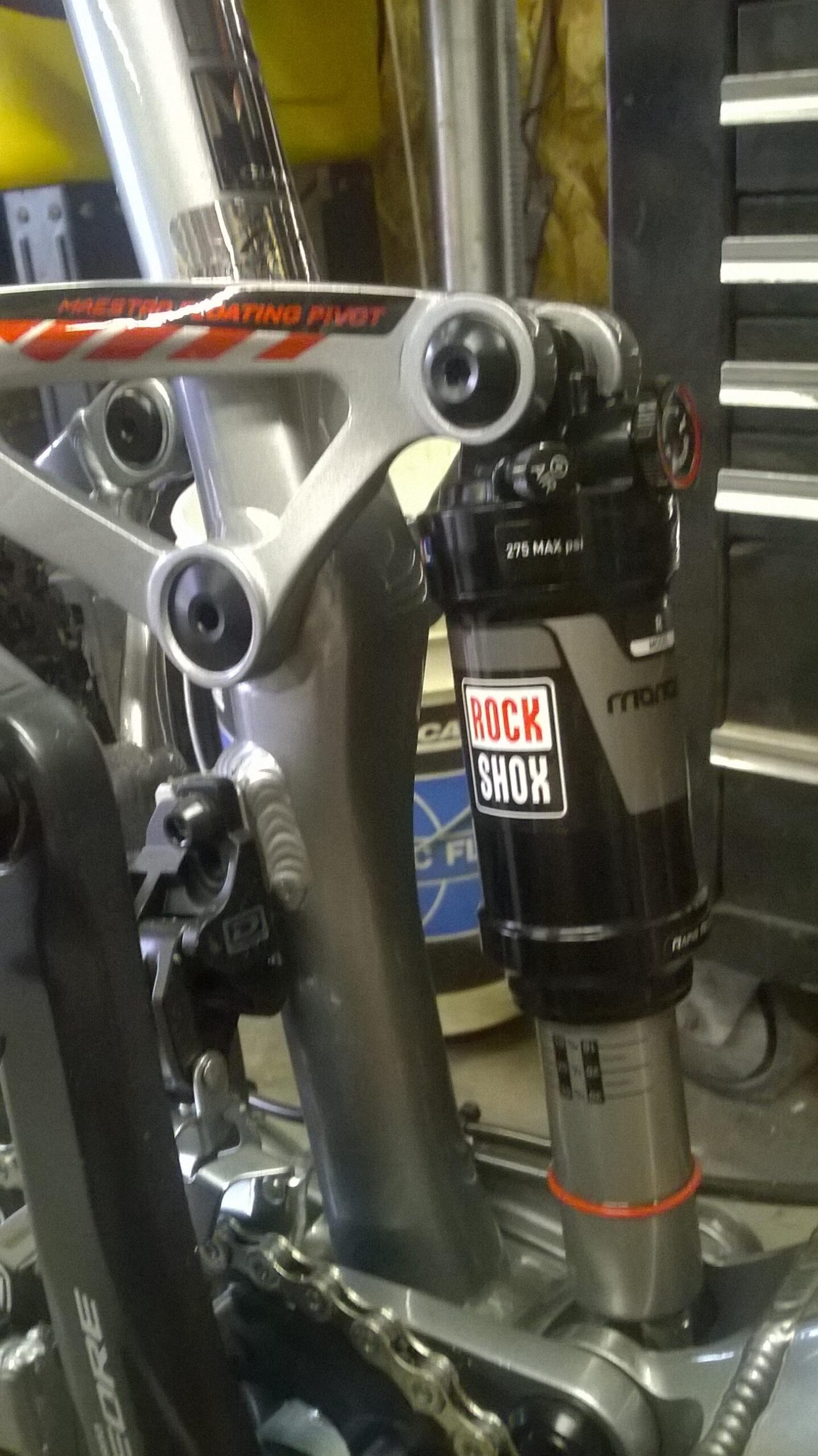 Giant Trance 27.5 3: Close-up image of a bicycle rear suspension system, featuring a RockShox shock absorber mounted on a silver frame with visible pivot points and chain. The background includes tools and equipment typically found in a workshop setting.