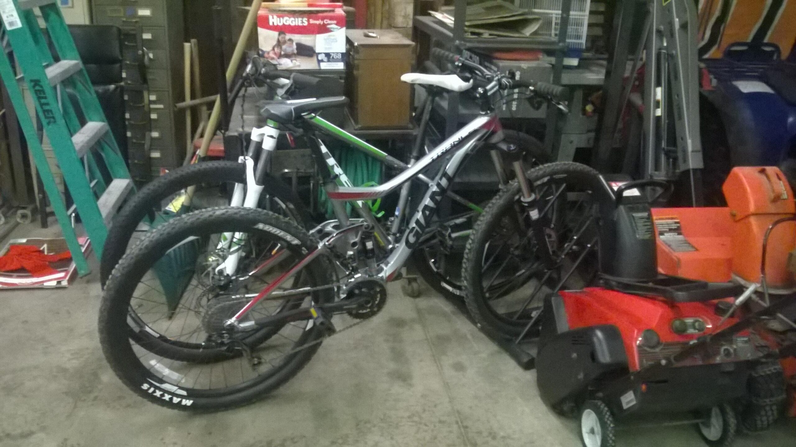 Giant Trance 27.5 3: Alt tag: A cluttered garage scene featuring two mountain bikes, one with a white frame and green accents, and the other in silver and red, alongside various tools and equipment including a snowblower, a ladder, and a broom.