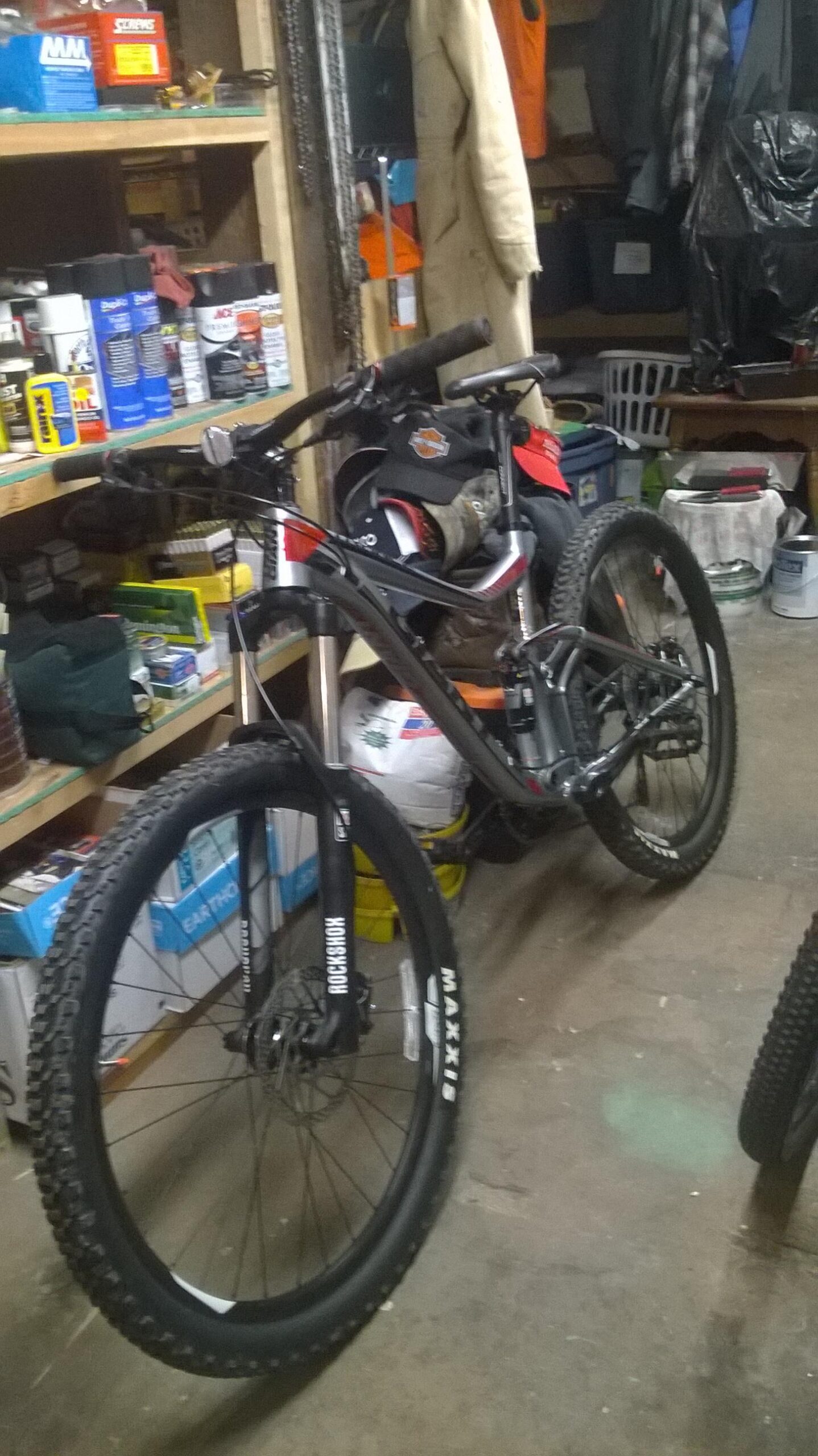 Giant Trance 27.5 3: A black and red mountain bike is propped up in a cluttered garage, surrounded by various tools and equipment. The bike features thick tires, a sturdy frame, and is positioned next to shelves filled with spray cans and other supplies. Various items, including a jacket and a hat, are visible in the background.