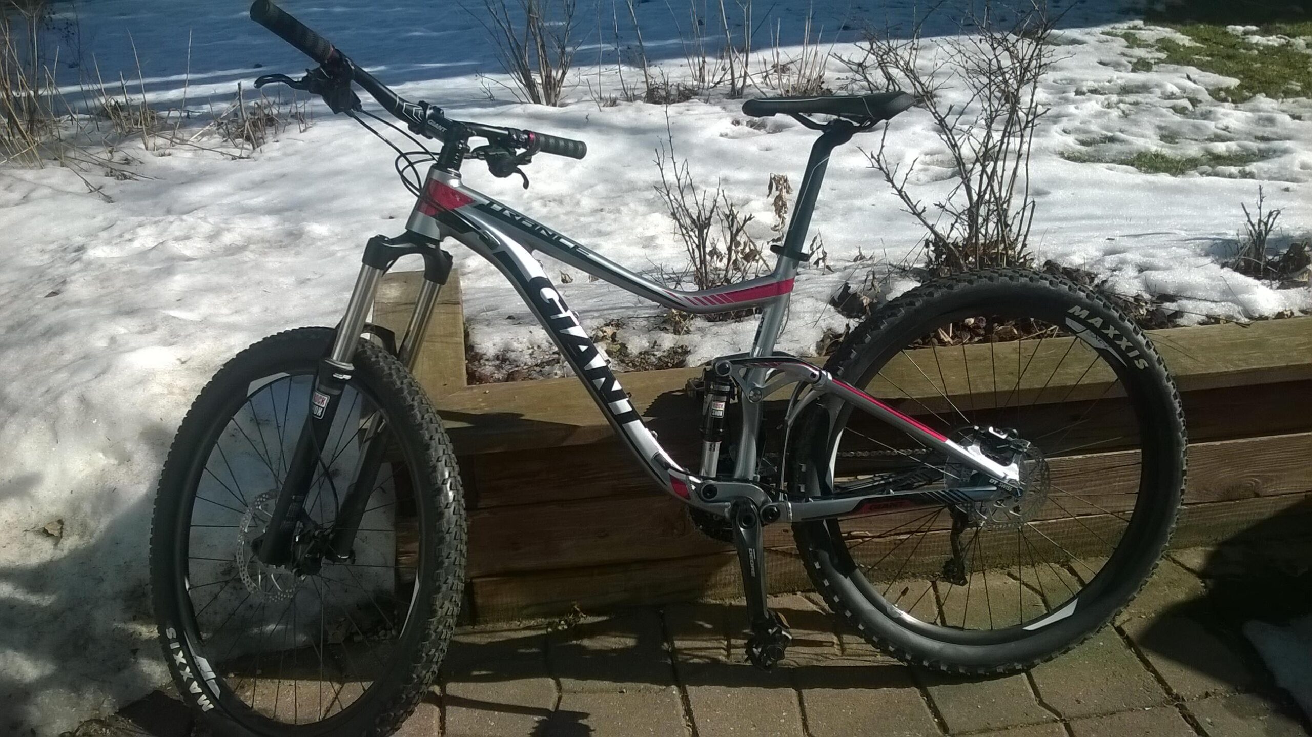 Giant Trance 27.5 3: A mountain bike leaning against a wooden planter, set against a background of melting snow and bare shrubs. The bike has a silver and black frame with red accents, featuring thick tires suitable for off-road riding.