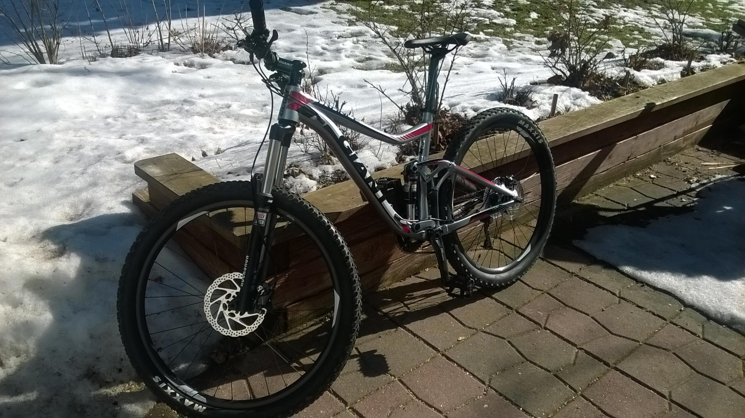 Giant Trance 27.5 3: A mountain bike with a silver and pink frame is resting against a wooden planter on a patio. Snow is visible on the ground, indicating a transition to spring. The bike features thick tires, a front suspension fork, and disc brakes. The background shows a partially snowy landscape with some greenery.