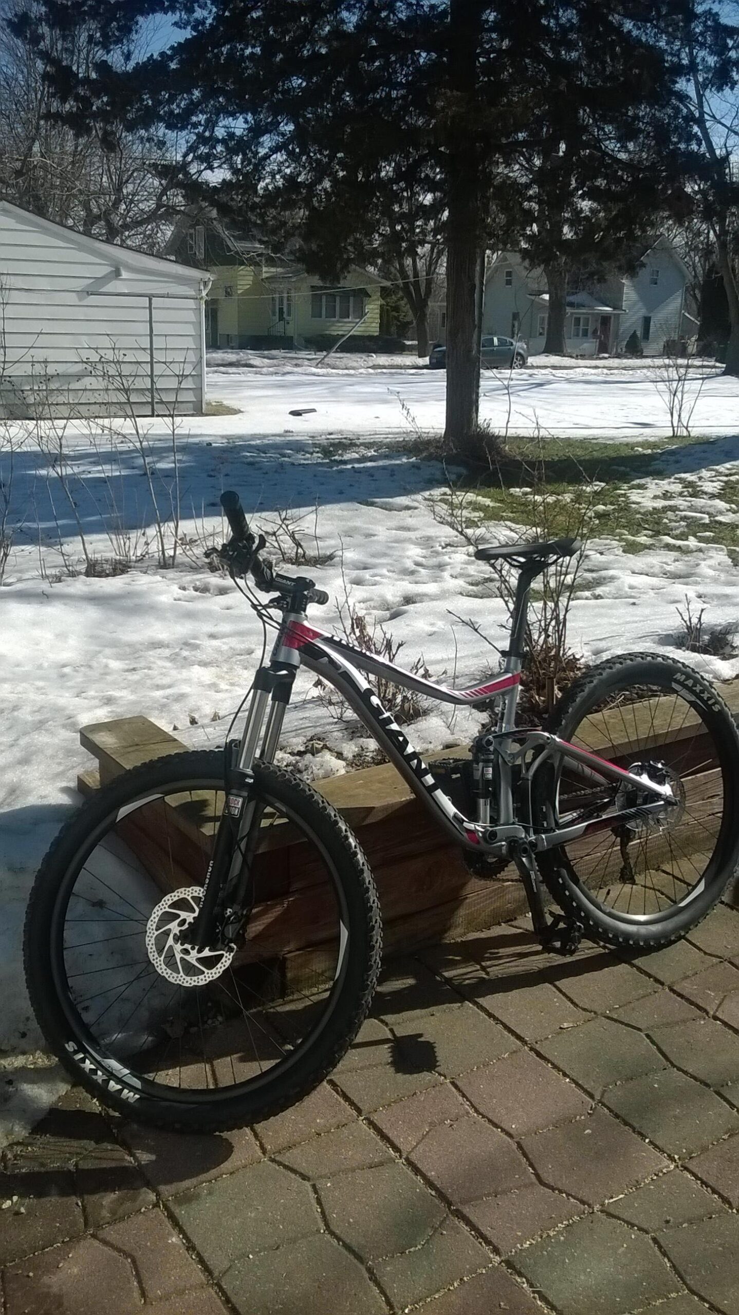 Giant Trance 27.5 3: A mountain bike with a silver and pink frame is resting on a wooden planter. In the background, there is a snowy yard and several houses visible under a clear blue sky. The ground is partially covered in snow, suggesting early spring weather.