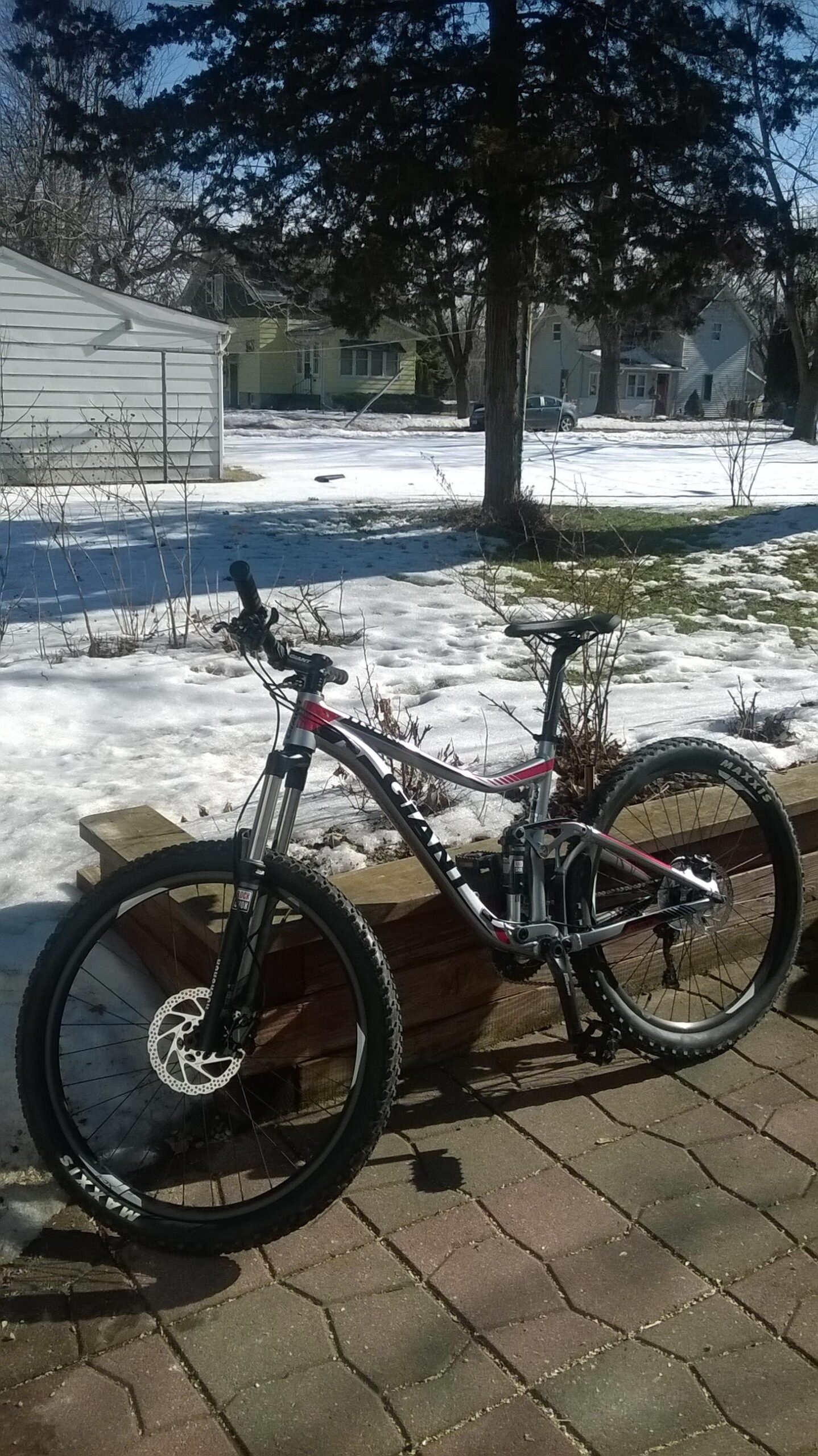 Giant Trance 27.5 3: A mountain bike is parked on a stone patio next to a wooden planter. In the background, there's a snowy yard with patches of grass and several houses visible. The sky is clear and blue, indicating a bright day.