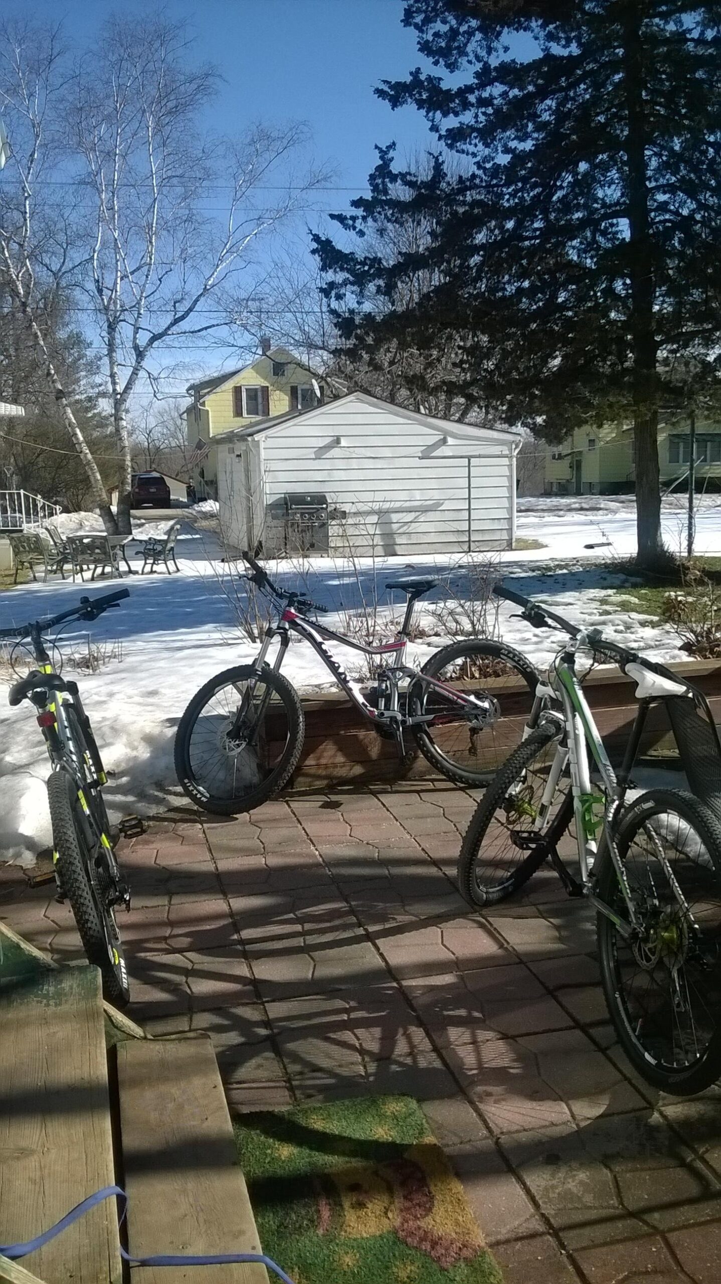 Giant Trance 27.5 3: Three bicycles are parked on a patio with a mix of snow and patches of grass visible in the background. A yellow house and a white shed are in the distance, under a clear blue sky. Some outdoor furniture can be seen in the background, along with a barbecue grill. The scene is set in a residential area, suggesting early spring conditions.