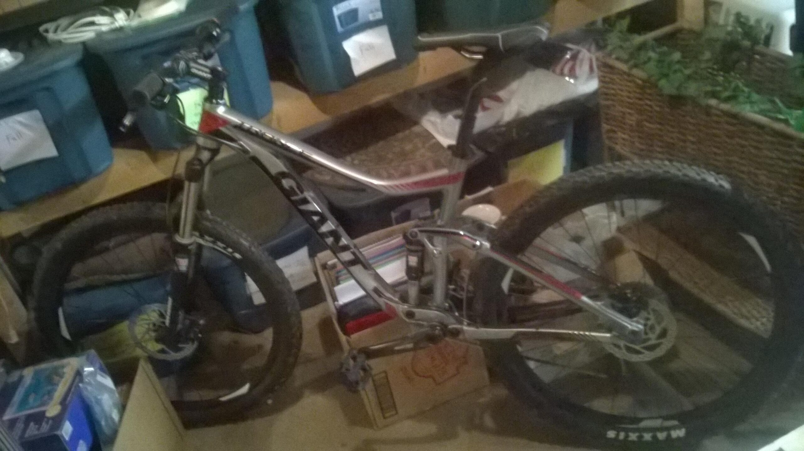 Giant Trance 27.5 3: A mountain bike leaning against storage boxes in a cluttered garage. The bike has a silver frame and thick, knobby tires, with the brand name "Giant" visible on the side. Various items, including storage bins and boxes, are scattered around the background.