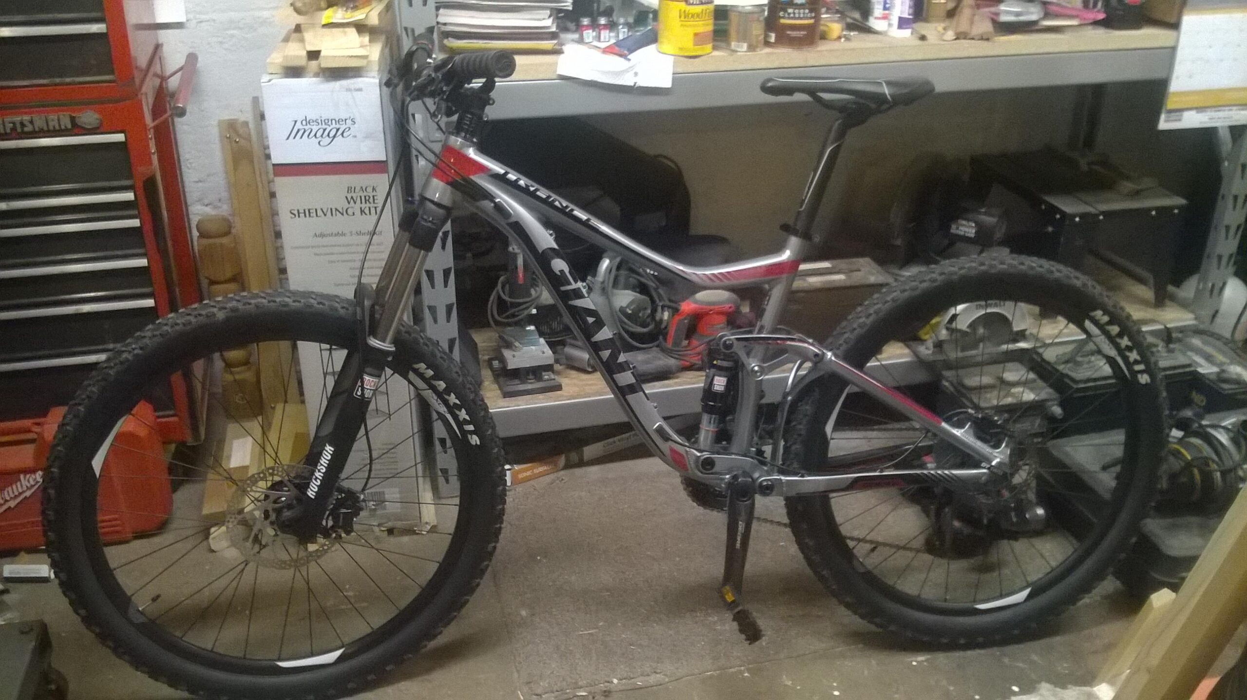 Giant Trance 27.5 3: A mountain bike with a sleek silver and red frame is positioned in a cluttered garage. The bike features thick, knobby tires, front suspension, and is resting on a concrete floor. Behind it, various tools and materials are visible on shelves, including a toolbox and wood pieces.
