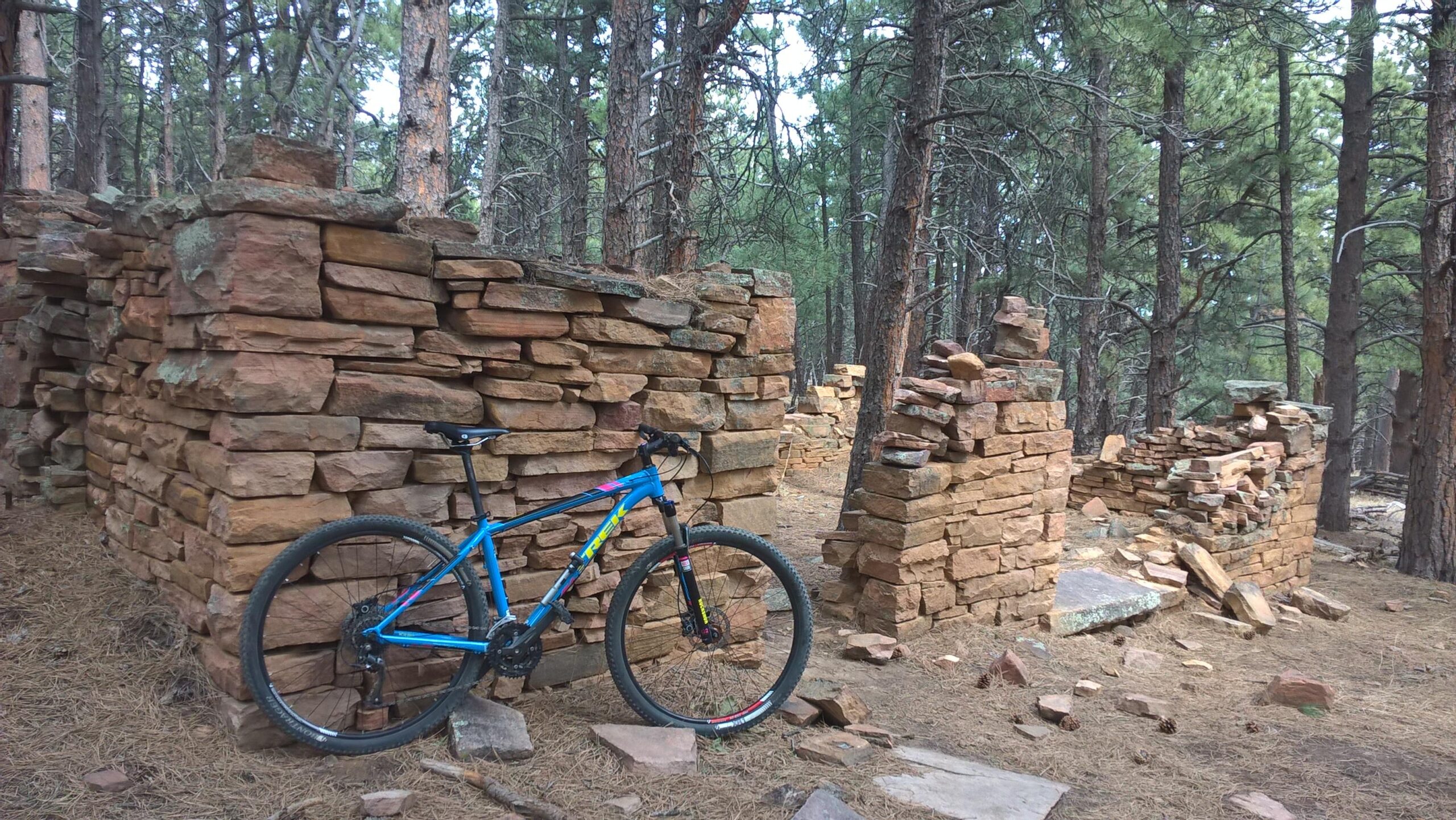 A blue mountain bike is leaning against a stone structure surrounded by tall, green pine trees in a forested area. The structure, made of reddish stones, appears to be in ruins, with some sections partially collapsed and scattered stones on the ground. Heil Valley Ranch mountain bike trail.