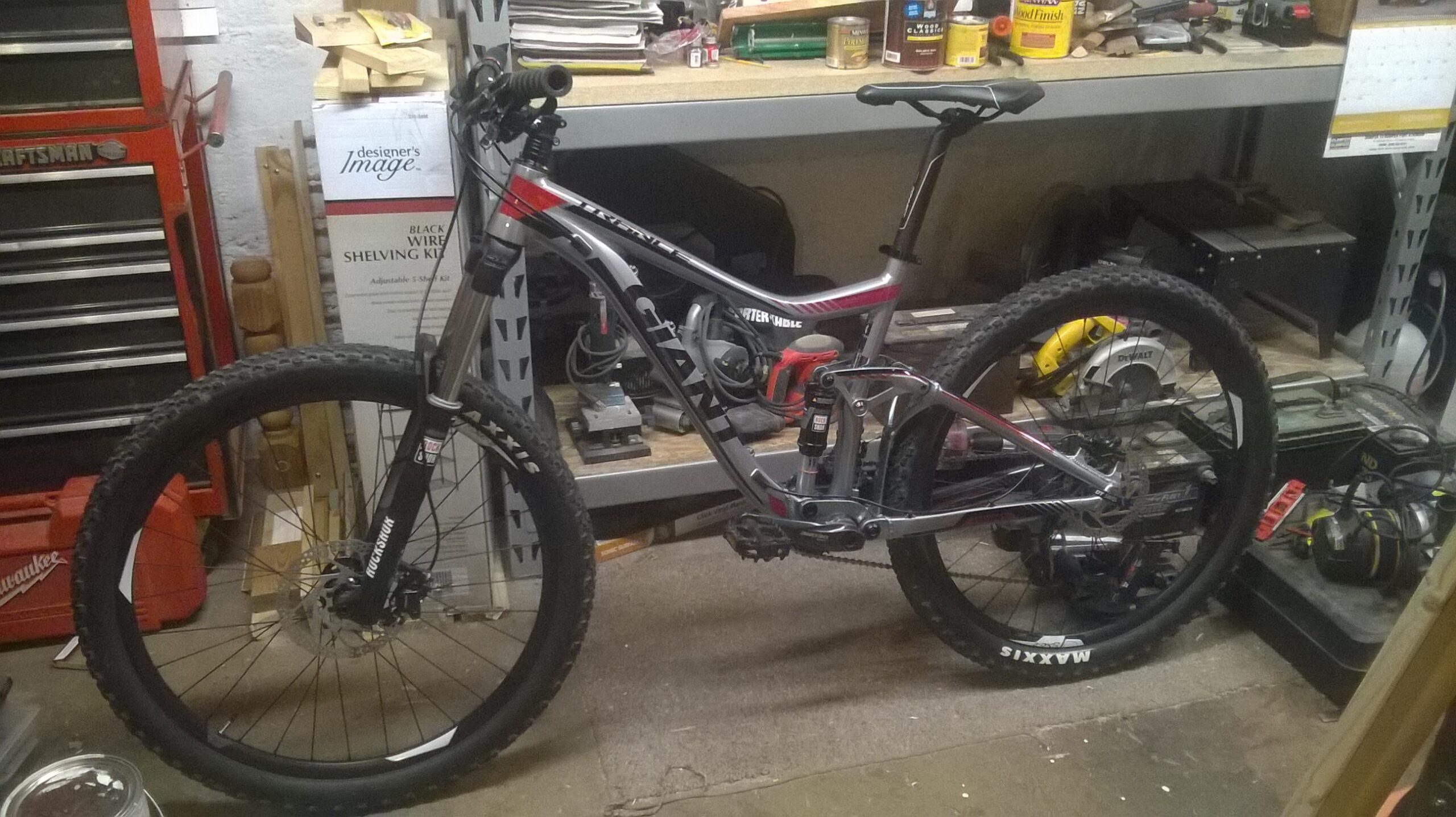 Giant Trance 27.5 3: A mountain bike with a sleek frame and knobby tires is positioned in a cluttered workshop. Various tools and equipment are visible in the background, including a toolbox, power tools, and shelves filled with supplies. The bike features front and rear suspension and a prominent brand name on its frame.