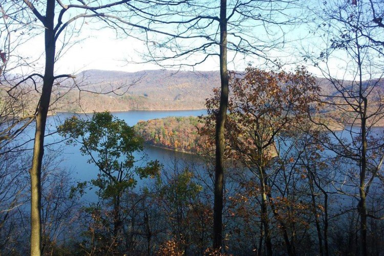 A picturesque view of a lake surrounded by trees, with an island in the center. The landscape showcases autumn foliage, reflecting vibrant colors against the serene blue water. In the background, rolling hills rise beneath a clear sky.