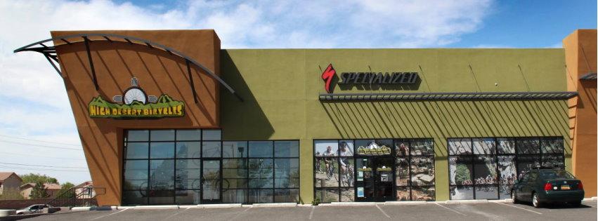 Image of a retail building with a modern design, featuring large glass windows and an angled roof. The exterior is painted in a bold green color with the logos of two businesses: one reads "Mike's Downtown Bicycles" on the left, and the other displays "Specialized" on the right. The surrounding area shows a parking lot with a car parked, under a clear blue sky.