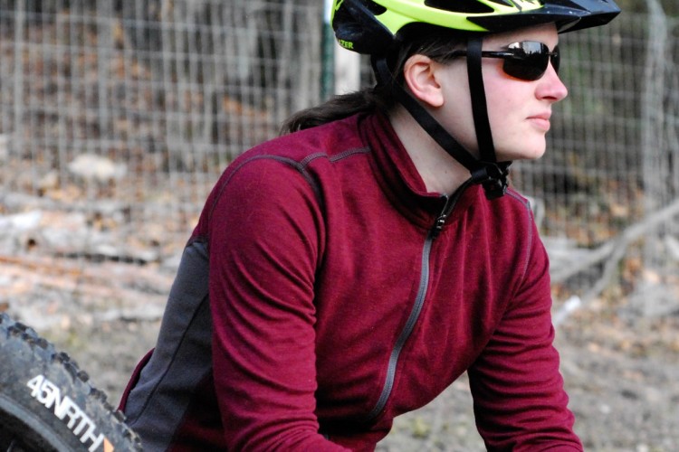 A person wearing a bright yellow bike helmet and sunglasses sits on the ground next to a mountain bike, dressed in a maroon long-sleeve jacket and black gloves. The background features blurred trees and a fence, suggesting an outdoor setting.