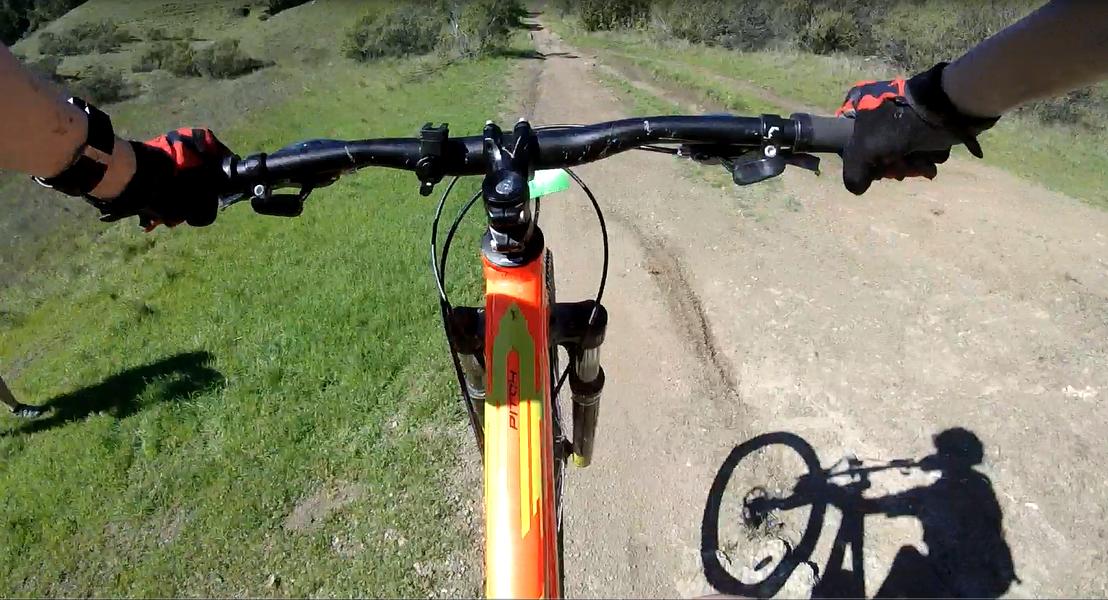 A close-up view of the handlebars of a mountain bike riding on a dirt path, with green grass and a sloping hillside in the background. The cyclist's hands are gripping the handlebars, and a shadow of the bike and rider can be seen on the ground. Camp Tamarancho mountain bike trail.