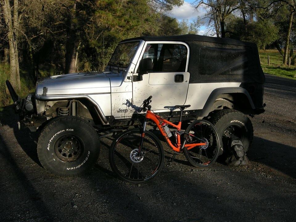 Trek Fuel EX 5 29: A silver Jeep Wrangler parked on a gravel path, with an orange mountain bike leaning against the vehicle's large tire. The background features trees and a partly cloudy sky.
