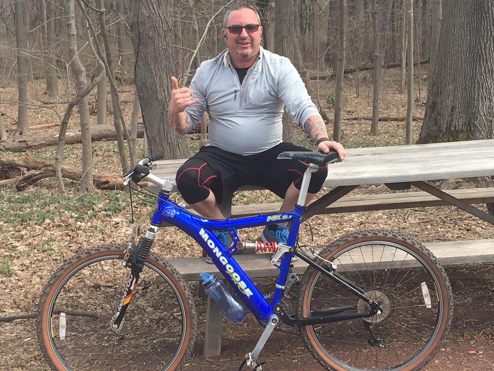 Mongoose MONGOOSE XR-PRO: A person sitting on a picnic table in a forested area, smiling and making a hand gesture. They are wearing a light gray athletic shirt and black shorts with padding. A blue mountain bike is parked next to them, featuring a water bottle attached to the frame. The background shows trees and fallen leaves, suggesting an outdoor setting in early spring.