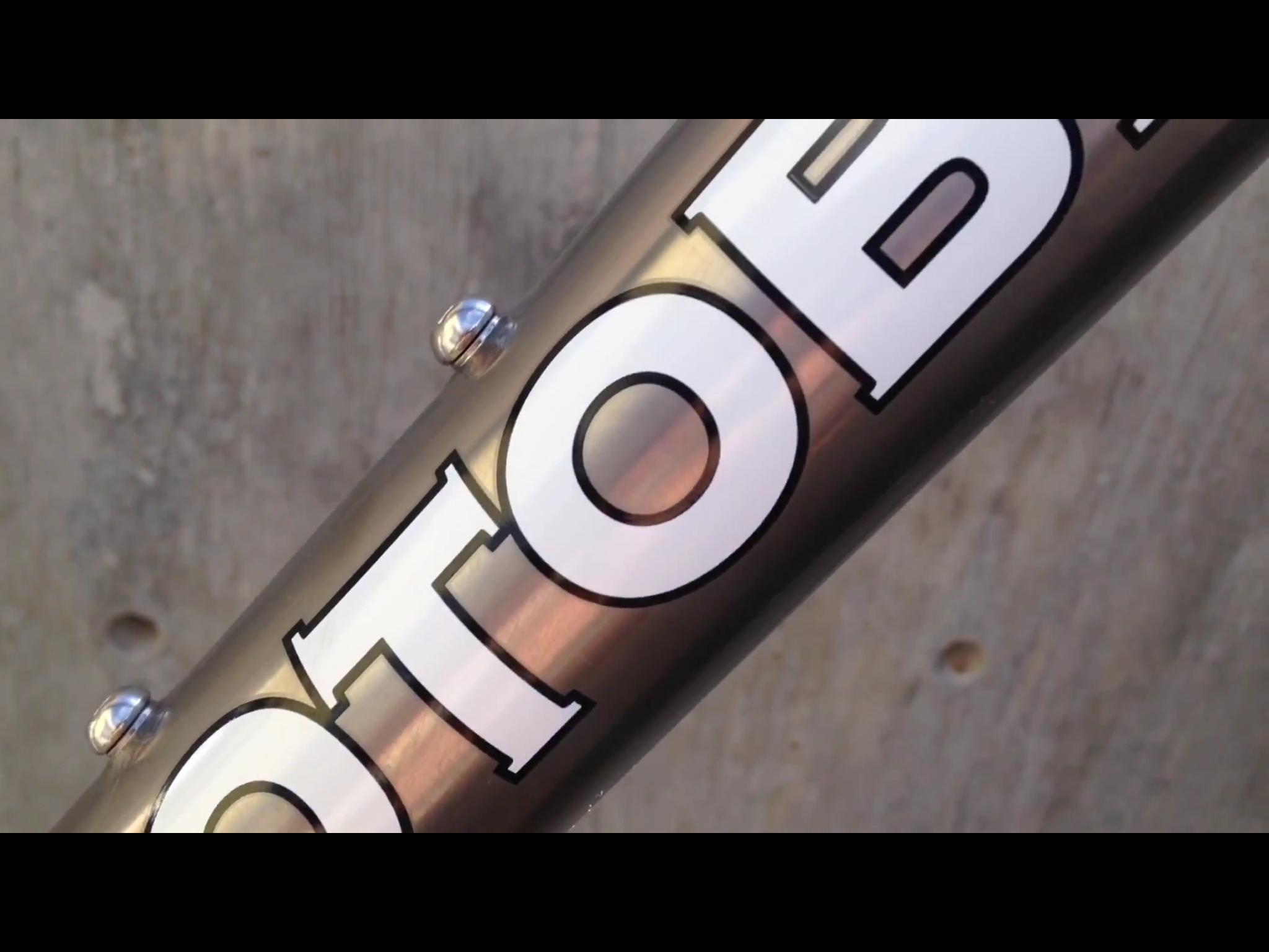 Motobecane Motobecane Bullet Titanium Fat bike: Close-up of a metallic bicycle frame showing the logo "OTOB" with a shiny silver bolt on the side, against a textured concrete background.