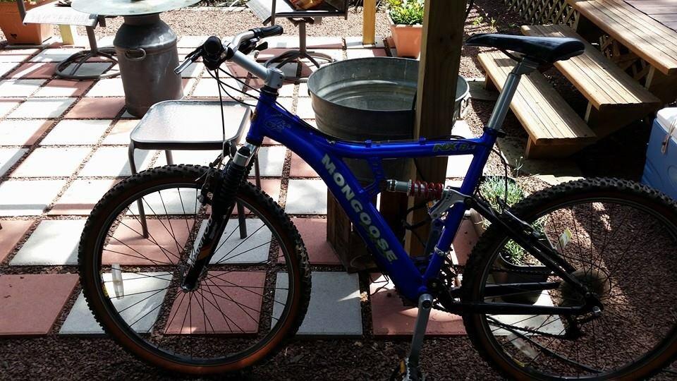 Mongoose MONGOOSE XR-PRO: A blue Mongoose mountain bike parked on a tiled patio, surrounded by outdoor furniture and potted plants. In the background, there's a metal bucket and wooden steps leading to a deck area. Sunlight illuminates the scene, highlighting the bike's details and the textured surfaces of the patio.