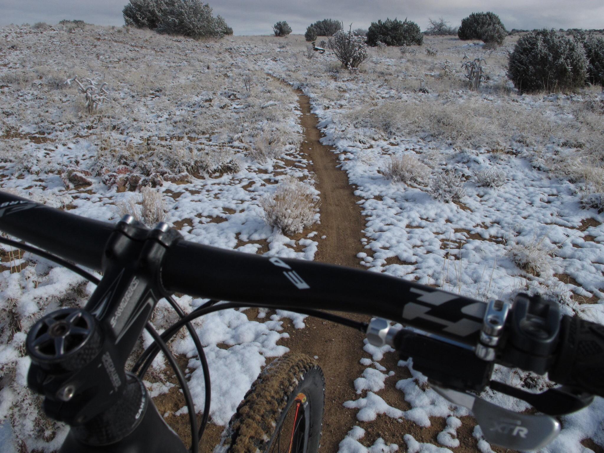 Close-up view of a mountain bike handlebar, with a snowy trail visible in the foreground. The path winds through a frosty landscape with patches of snow covering the ground and sparse vegetation, under a cloudy sky. Parkway Fatbike trail mountain bike trail.