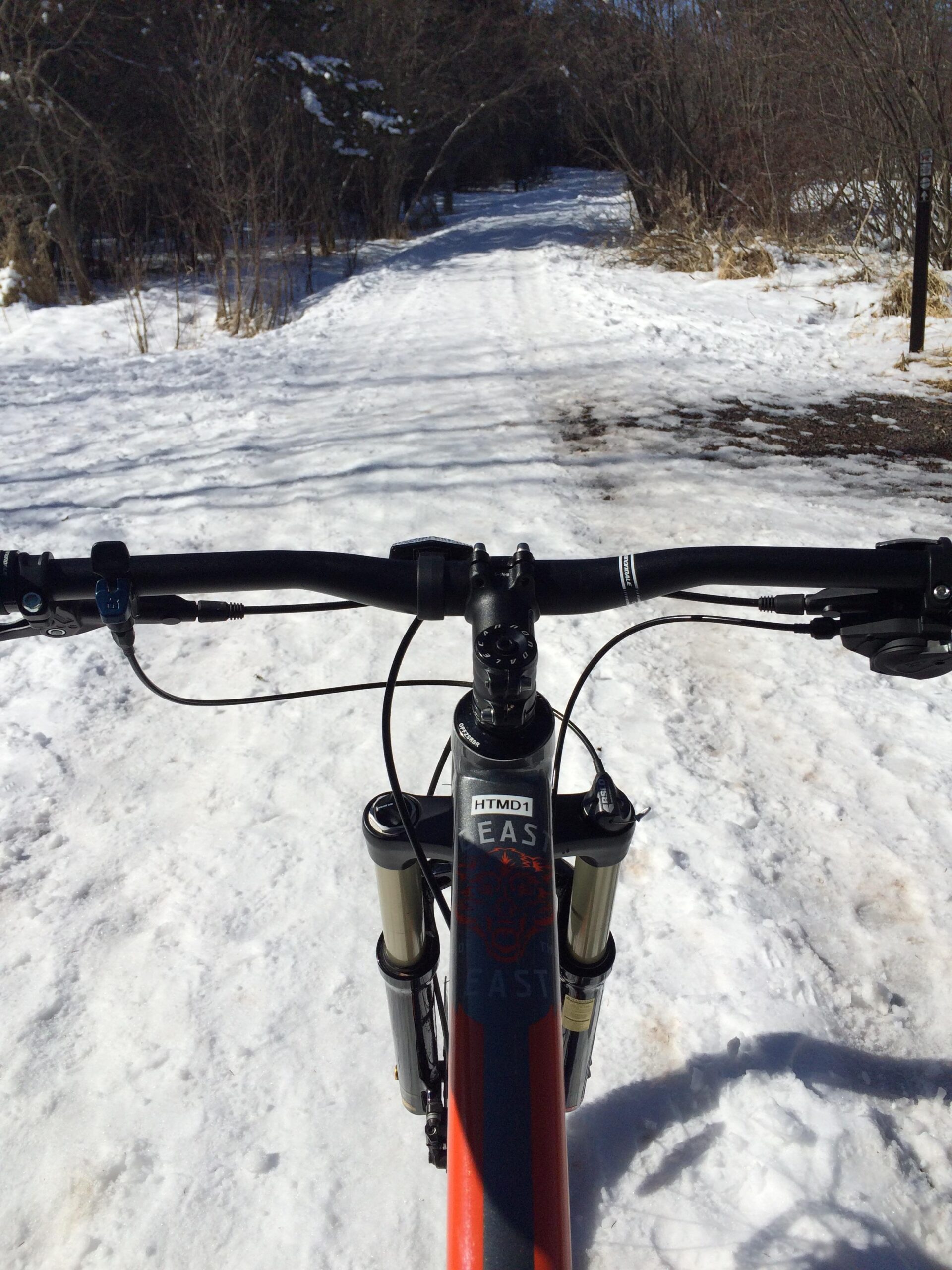 Cannondale Beast of the East: View from the handlebars of a mountain bike on a snowy path, with trees lining the sides and a clear blue sky above. The bike frame is mainly black with red accents, and the path is partially covered in snow, indicating winter conditions.