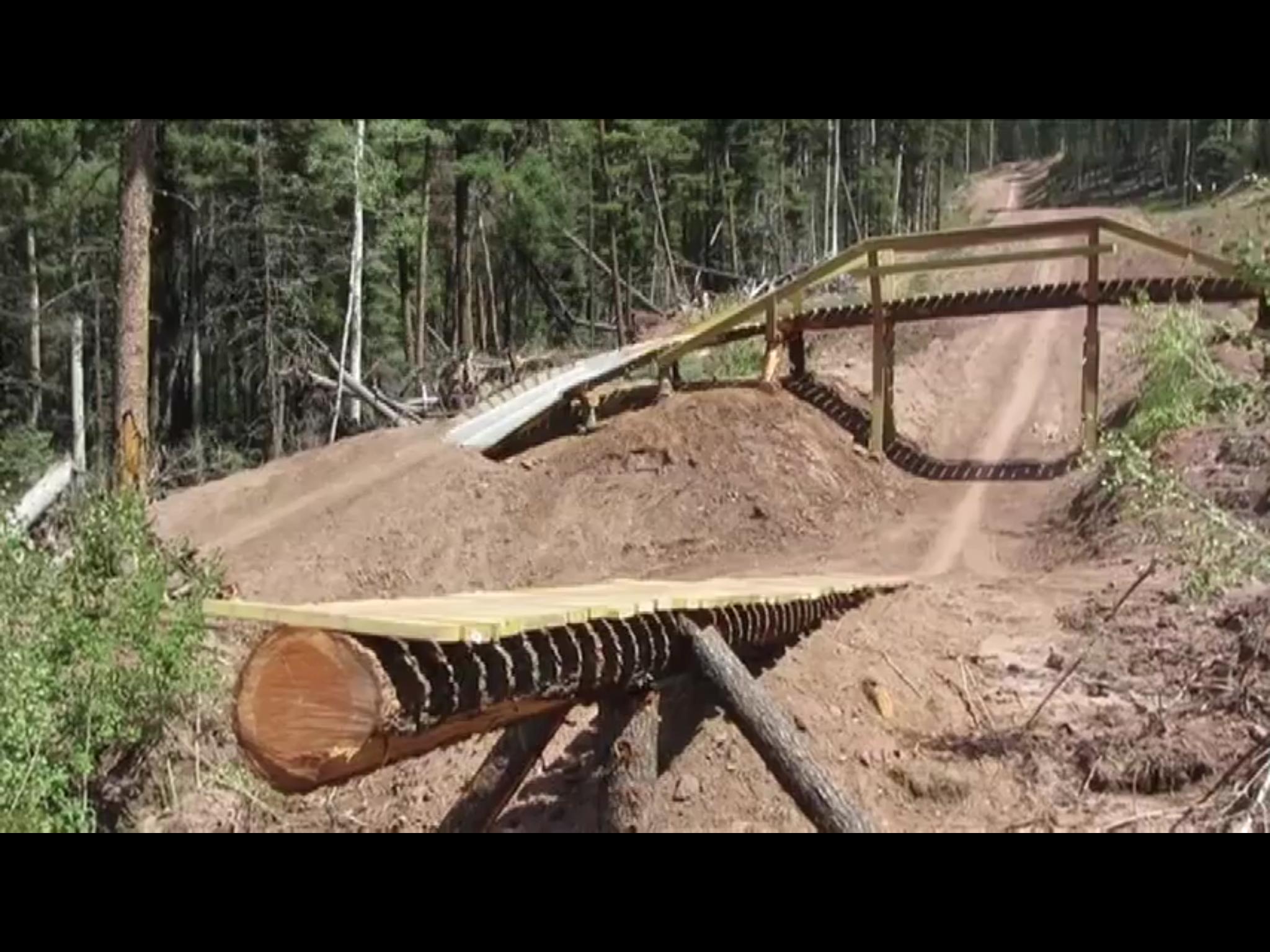 A wooden bike ramp built over a dirt mound in a forested area, with a winding dirt trail visible in the background. The ramp includes a curved section and is supported by logs and wooden beams. Surrounding trees and underbrush indicate a natural outdoor setting. Angel Fire Bike Park mountain bike trail.