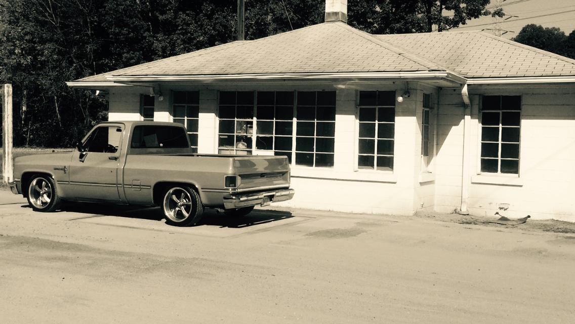 A vintage pickup truck parked outside a small white building with large windows. The scene has a monochromatic filter, giving it a classic feel. Trees are visible in the background, indicating a natural setting. Warrior Creek mountain bike trail.
