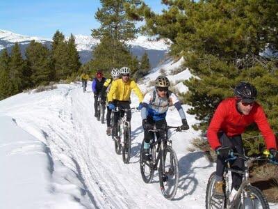 A group of six cyclists riding mountain bikes on a snowy trail, surrounded by trees and mountains in the background. The cyclists are wearing colorful winter cycling gear, with some snow visible on the path. Los Robles Trail (Western Section) mountain bike trail.