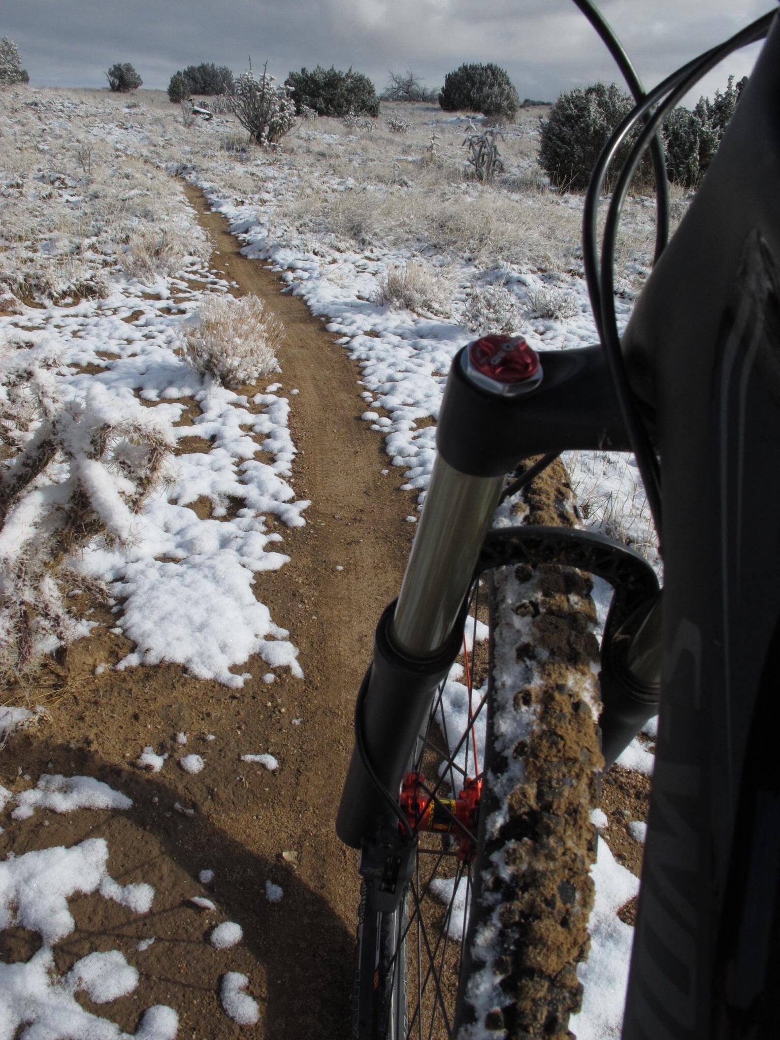 A close-up view of a mountain bike resting on a snowy trail. The path is a mix of dirt and snow, surrounded by sparse vegetation and low shrubs under a cloudy sky. The bike's front wheel and suspension are prominently featured, highlighting the contrast between the bike and the winter landscape. Parkway Fatbike trail mountain bike trail.