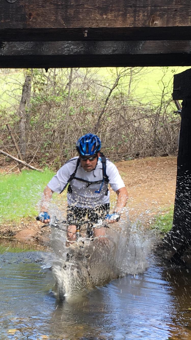 A mountain biker riding through a shallow stream, creating splashes of water. He is wearing a blue helmet, sunglasses, and a white shirt, with a rugged background of greenery and trees. The scene captures the adventurous spirit of outdoor cycling. El Dorado Trail mountain bike trail.