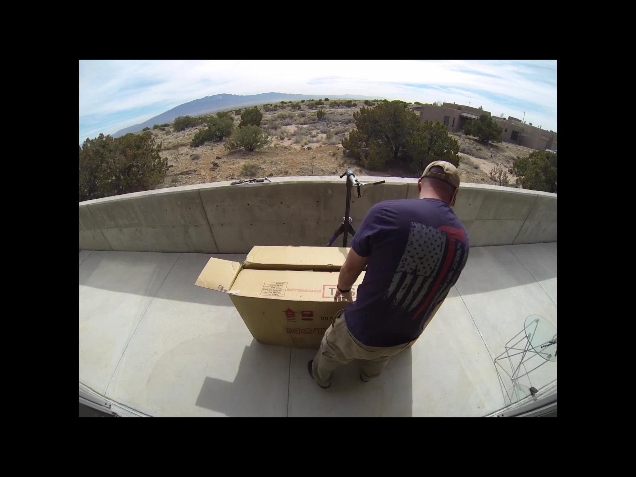 Motobecane Motobecane Bullet Titanium Fat bike: A person stands on a balcony, facing away from the camera, as they prepare to open a large cardboard box. The background showcases a desert landscape with scattered shrubs and mountains in the distance under a partly cloudy sky. A scooter and a metal chair are visible nearby.