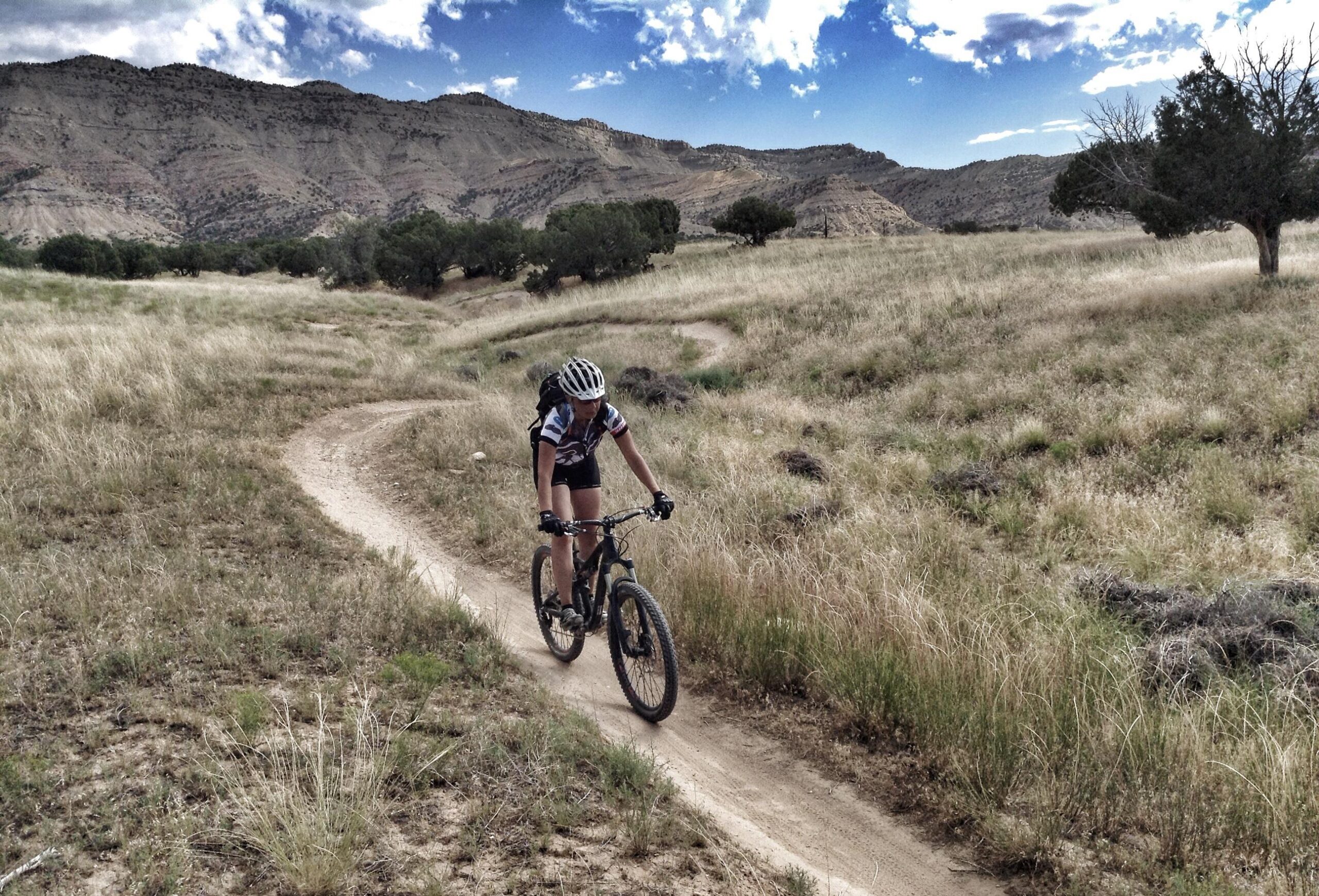 A person riding a mountain bike on a winding dirt trail through a grassy landscape with sparse trees, set against a backdrop of rugged hills and a partly cloudy sky. 18 Road Trails / North Fruita Desert mountain bike trail.