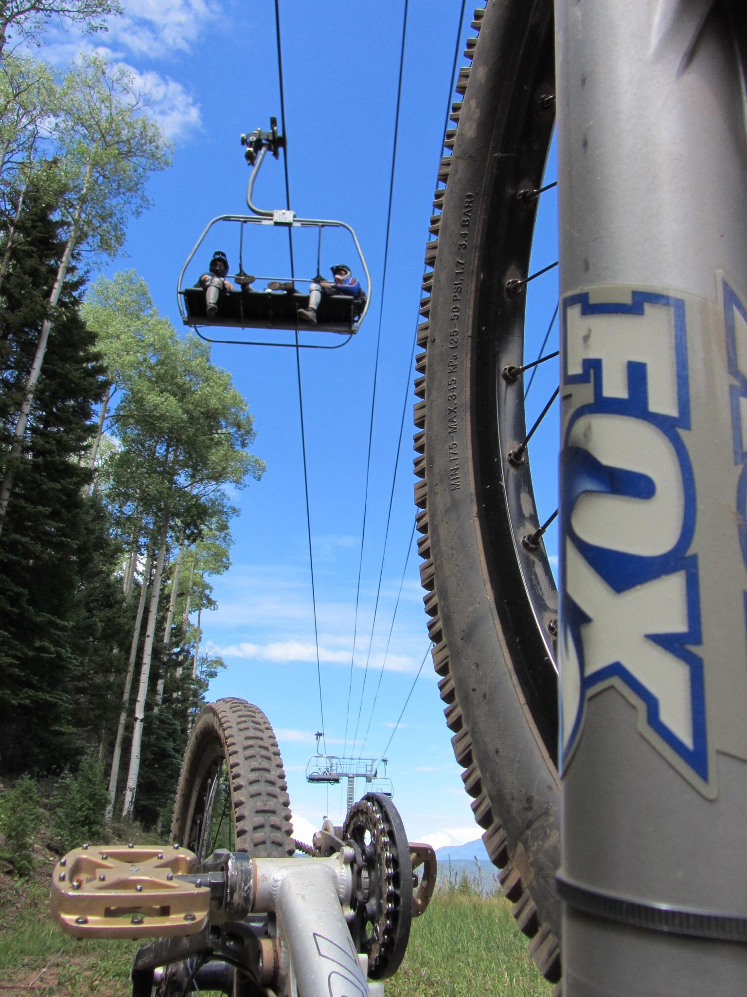 A close-up view of a mountain bike's wheel and pedal, with a ski lift in the background carrying two riders. The scene is set against a bright blue sky dotted with clouds, and green trees are visible on either side of the bike. Angel Fire Bike Park mountain bike trail.